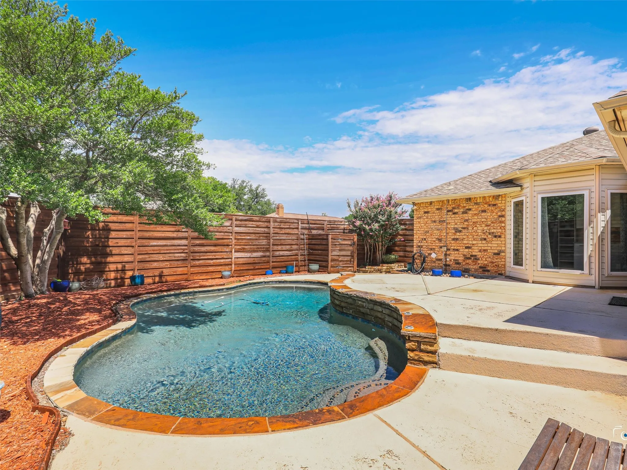 Curved swimming pool with stone coping, surrounded by a concrete patio and flower beds.