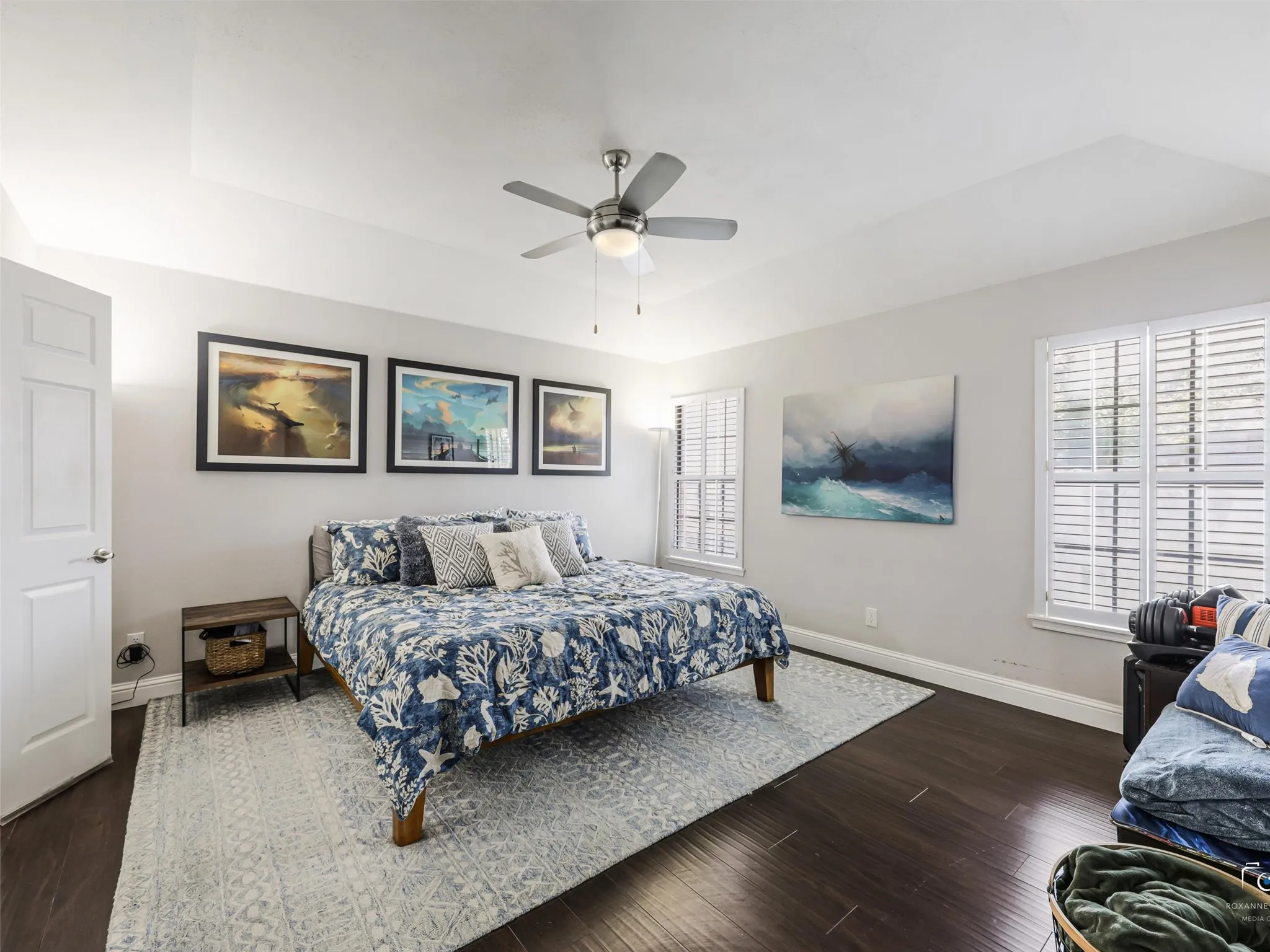 The same dark wood floors continue into the primary bedroom, along with a ceiling fan and windows with shutters.