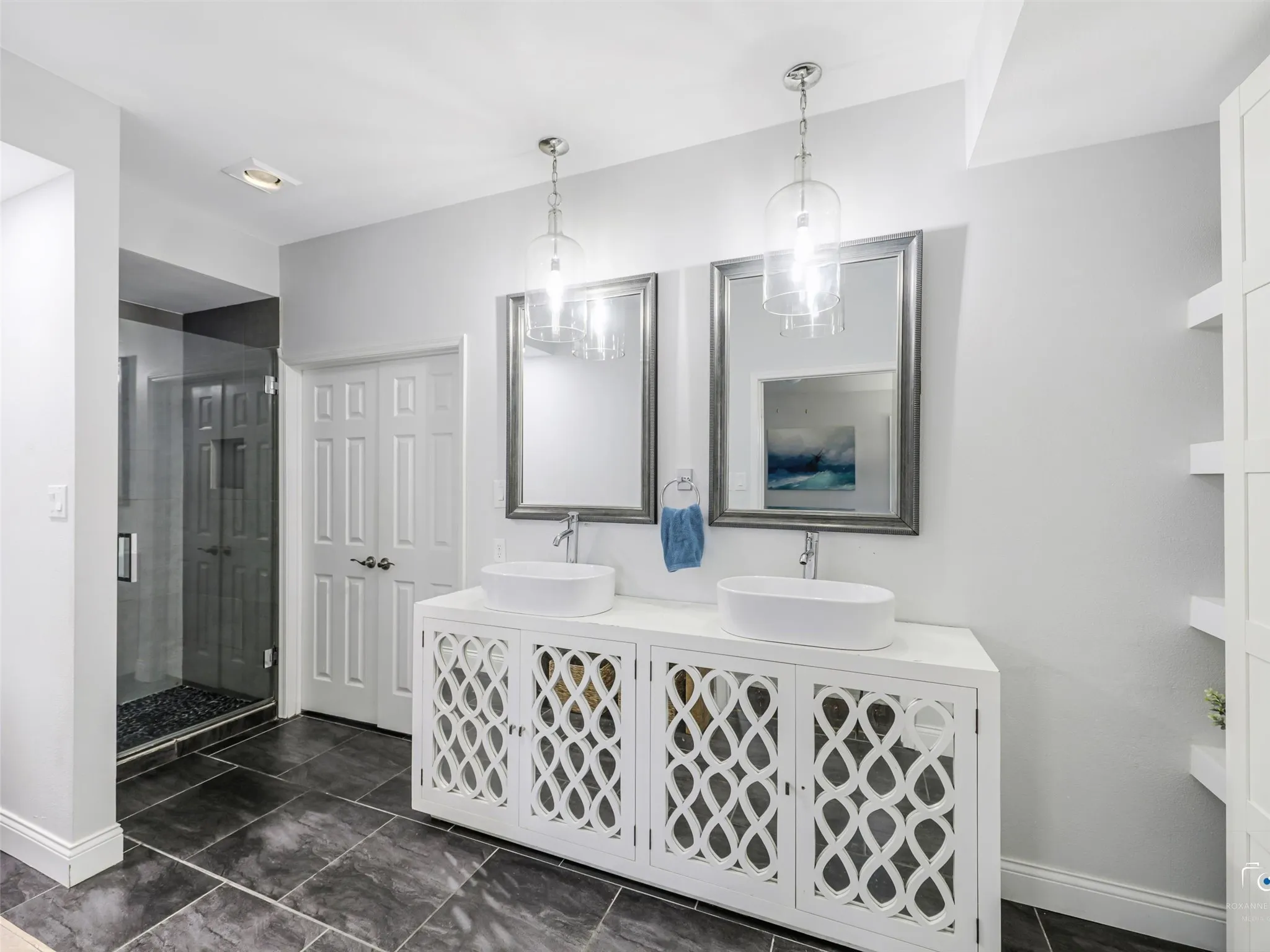 Contemporary bathroom featuring a double vanity with vessel sinks and decorative cabinet fronts, tile floors and a large walk-in shower.  The spacious primary closet is beyond the double doors.