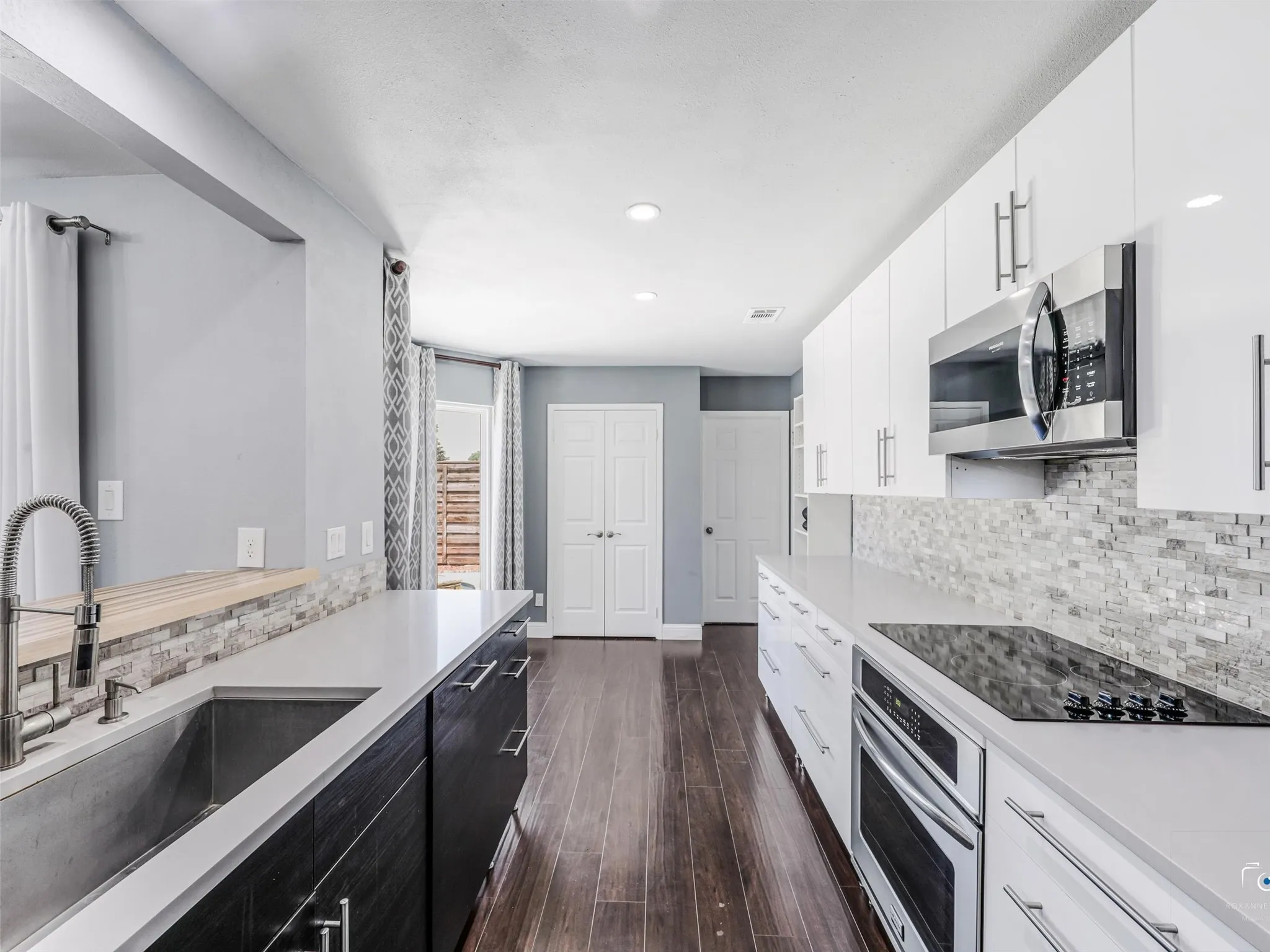 Modern kitchen featuring wood-finish flooring, white flat-panel upper cabinetry, dark flat-panel lower cabinetry, light-toned countertops, and a full-height tile backsplash.  The pantry is at the left double doors and the laundry room is beyond the door to the right. Note teh large sink!