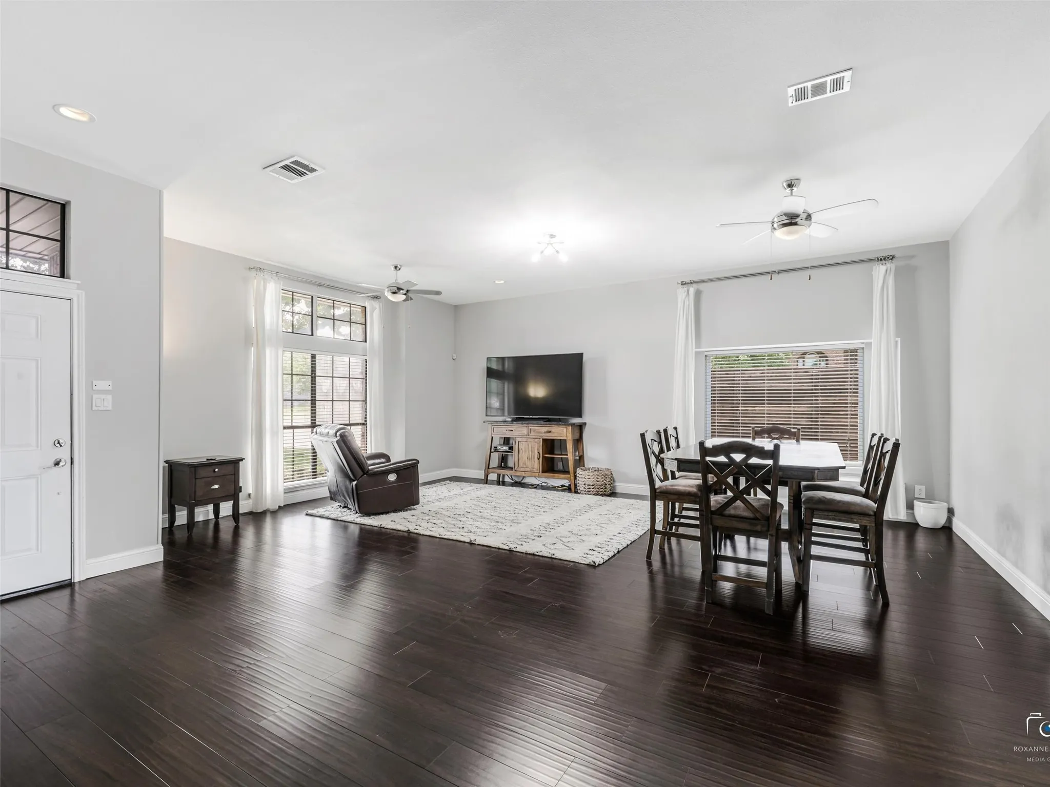 Open-concept living space features a living area and dining area with wood flooring, light gray walls, recessed lighting, two multiple ceiling fans and large windows providing ample natural light.