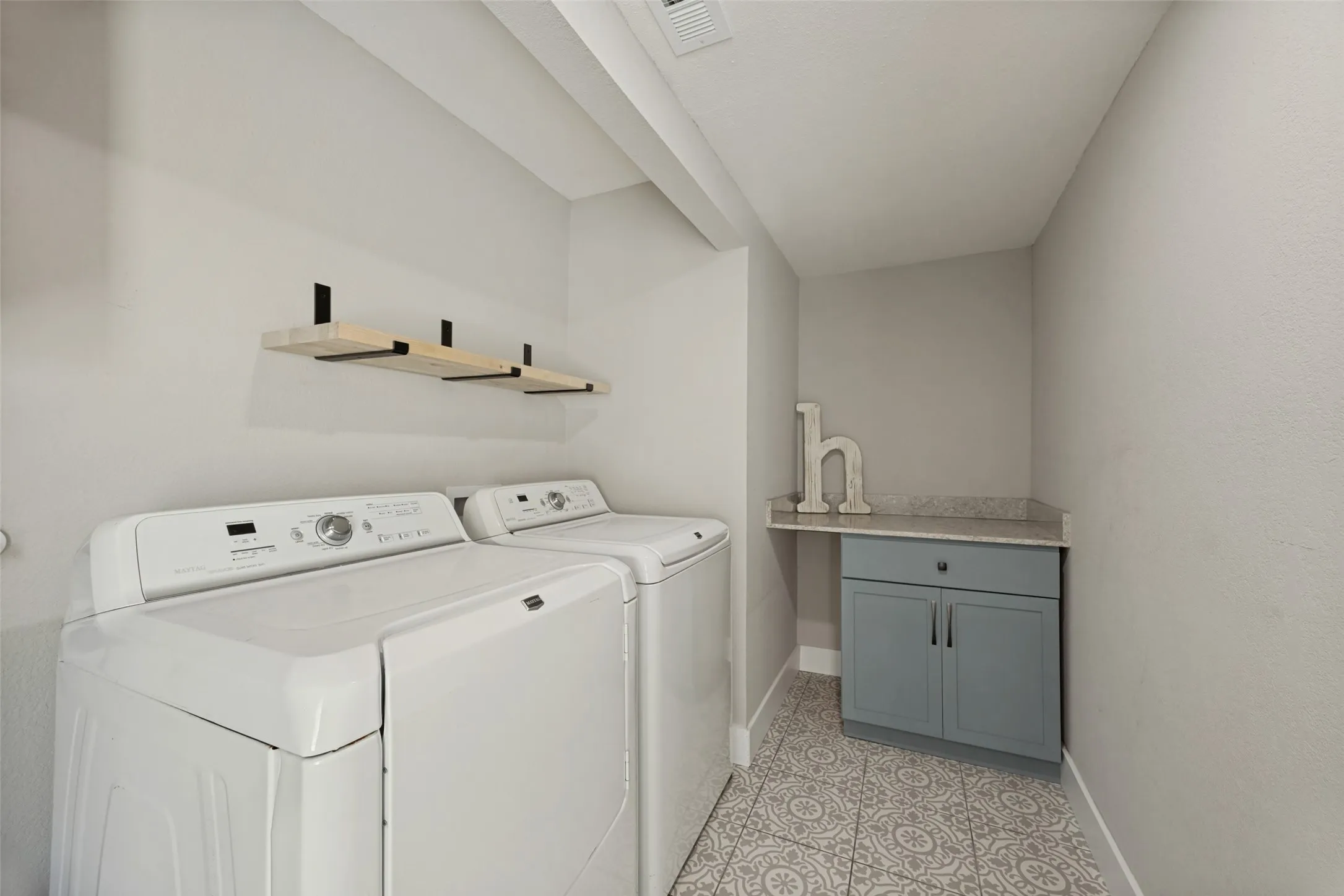 Laundry area featuring patterned tile flooring, wall-mounted wood shelves with dark brackets, and a built-in cabinet with a countertop