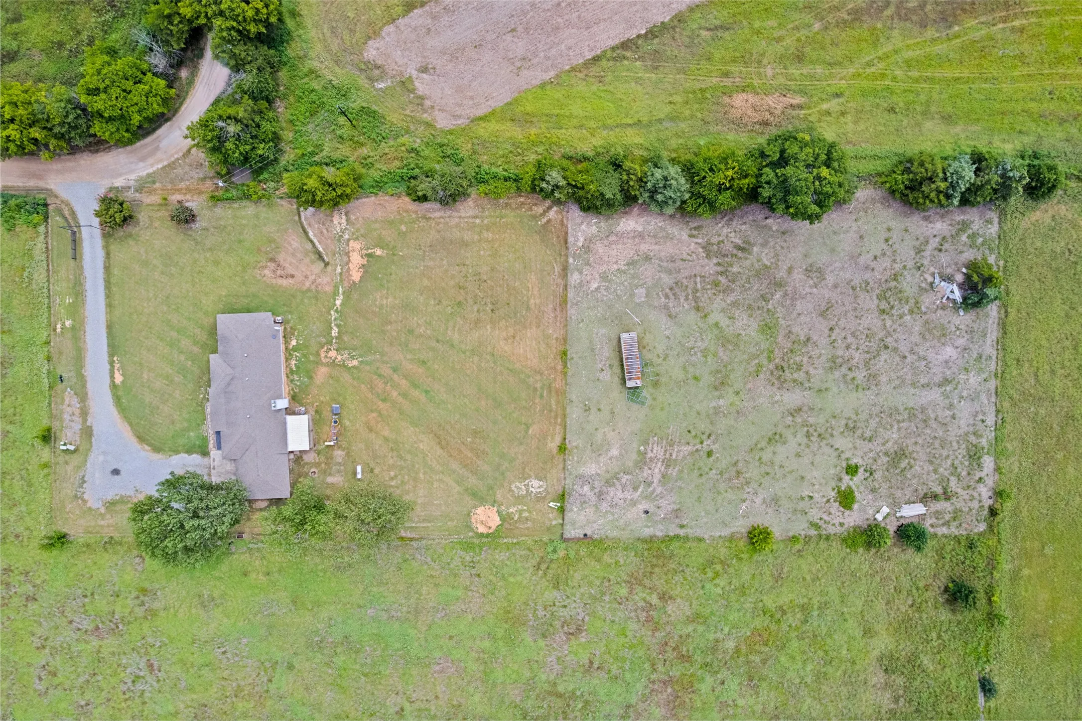 Expansive property featuring a single-story residence with a gray shingle roof, a long asphalt driveway, surrounding green spaces, and a separate fenced area with a small rectangular structure