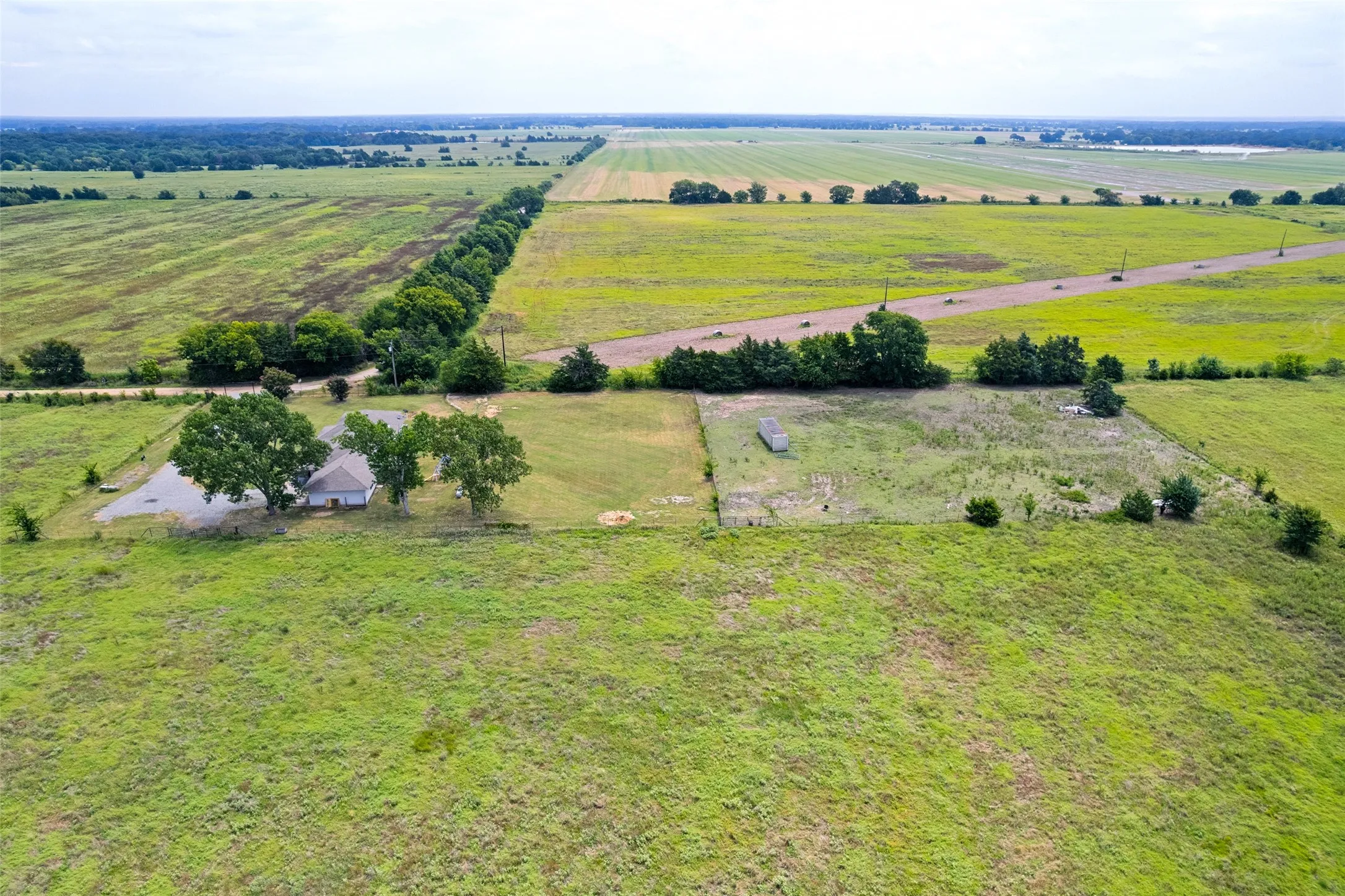 Expansive green acreage featuring a single-story residence with a light-colored roof, multiple mature trees, and a paved driveway