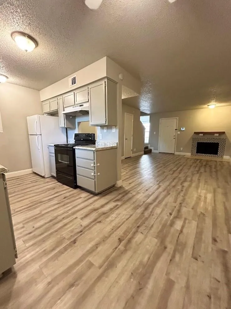 Open-concept living area featuring wood-finish flooring, a kitchen with light-toned cabinetry and black appliances, a tiled fireplace, and ceiling-mounted lighting fixtures