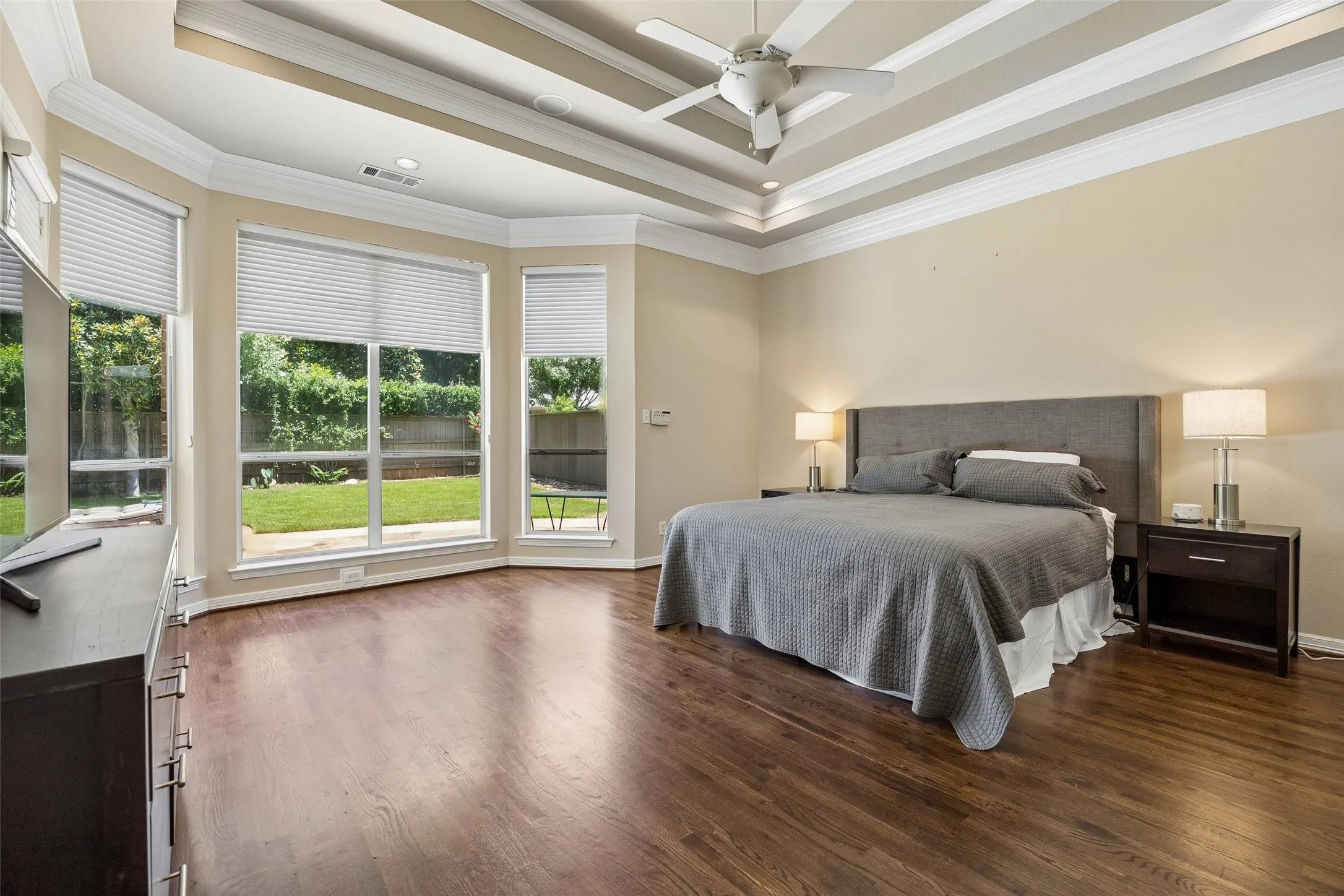 Expansive room featuring rich wood-finish flooring, a coffered ceiling with recessed lighting and a ceiling fan, and large bay windows with blinds overlooking a private backyard
