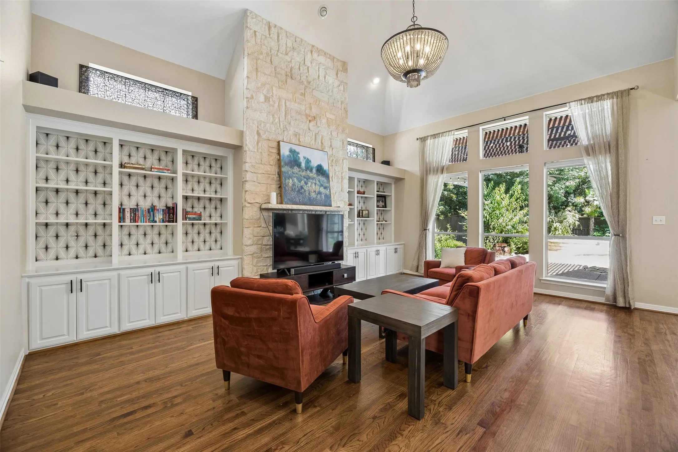 Spacious living area featuring high ceilings, wood-finish flooring, a stone-clad fireplace, built-in shelving, and large windows