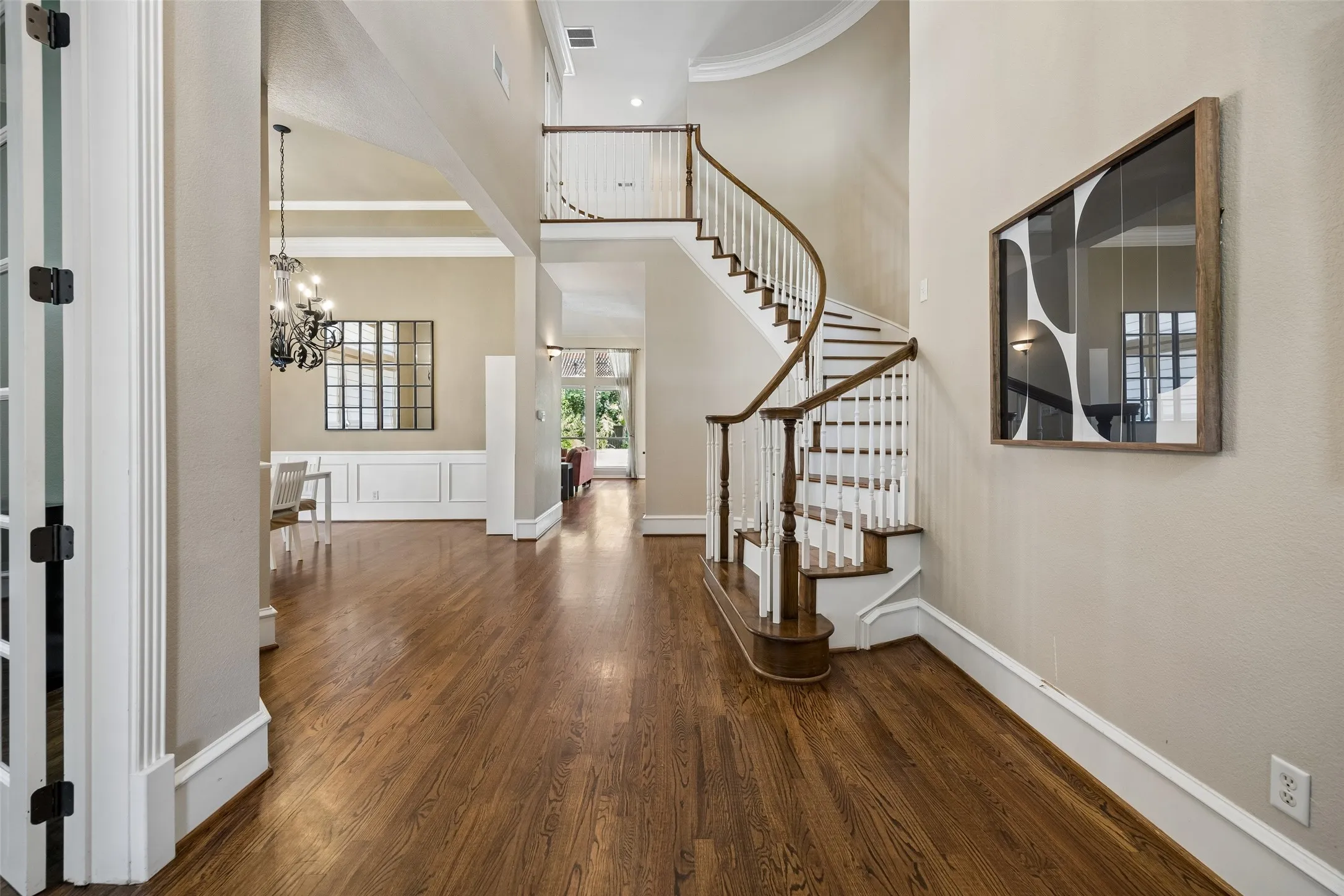 Grand foyer featuring wood-finish flooring, a curved staircase with white balusters and wood handrails, tall ceilings, and classic white wainscoting in the adjacent room