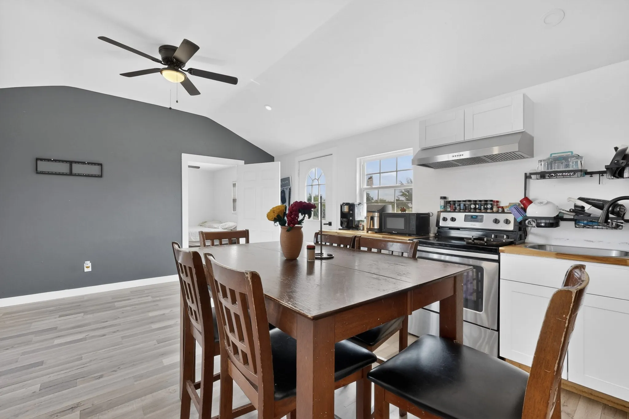 Open concept living area featuring wood-finish flooring, a vaulted ceiling with a ceiling fan, and a kitchen with white cabinetry, stainless steel appliances, and a window