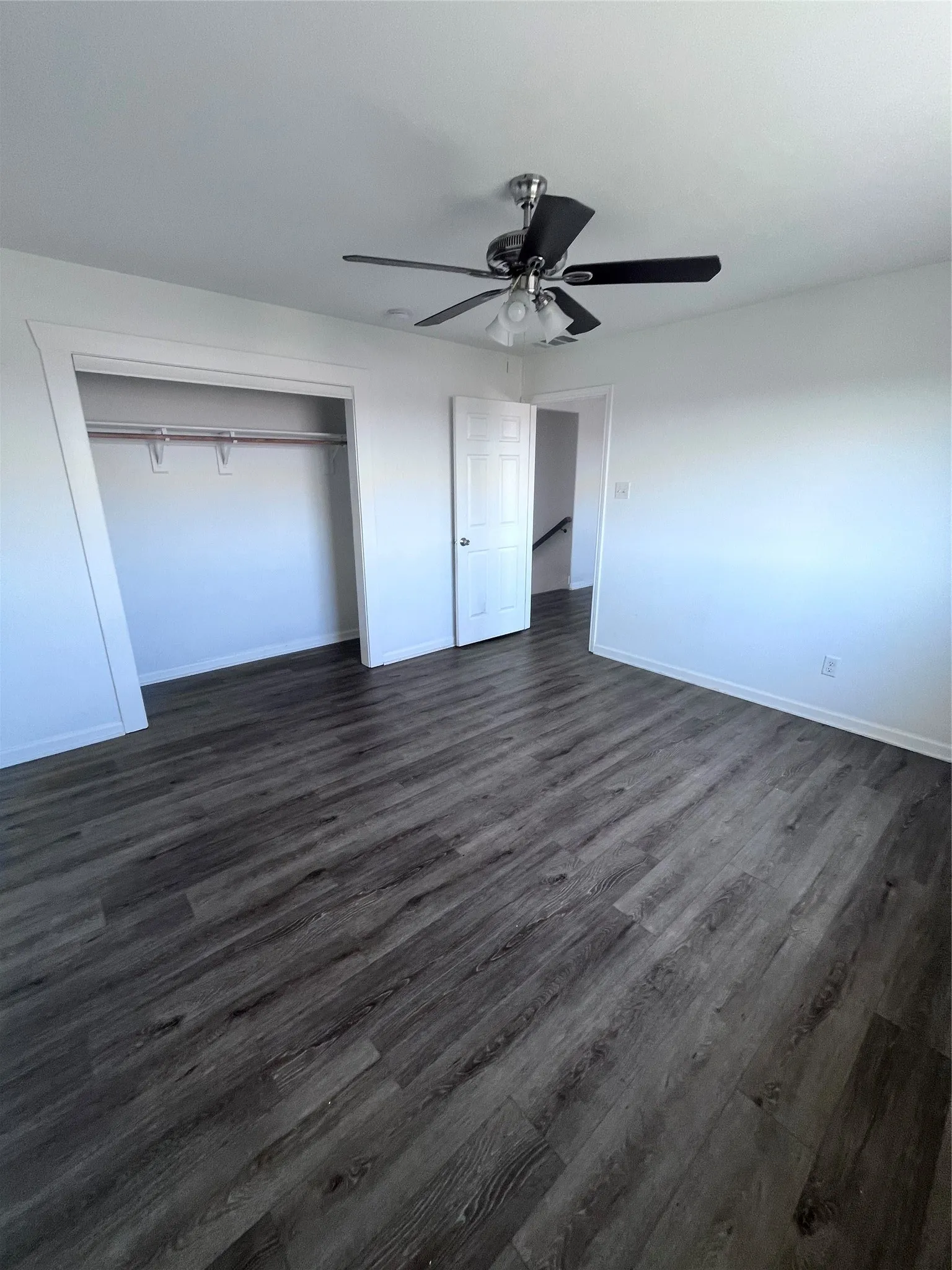 Spacious room featuring dark wood-finish flooring, a ceiling fan with integrated lighting, white walls, and an open closet space with a hanging rod