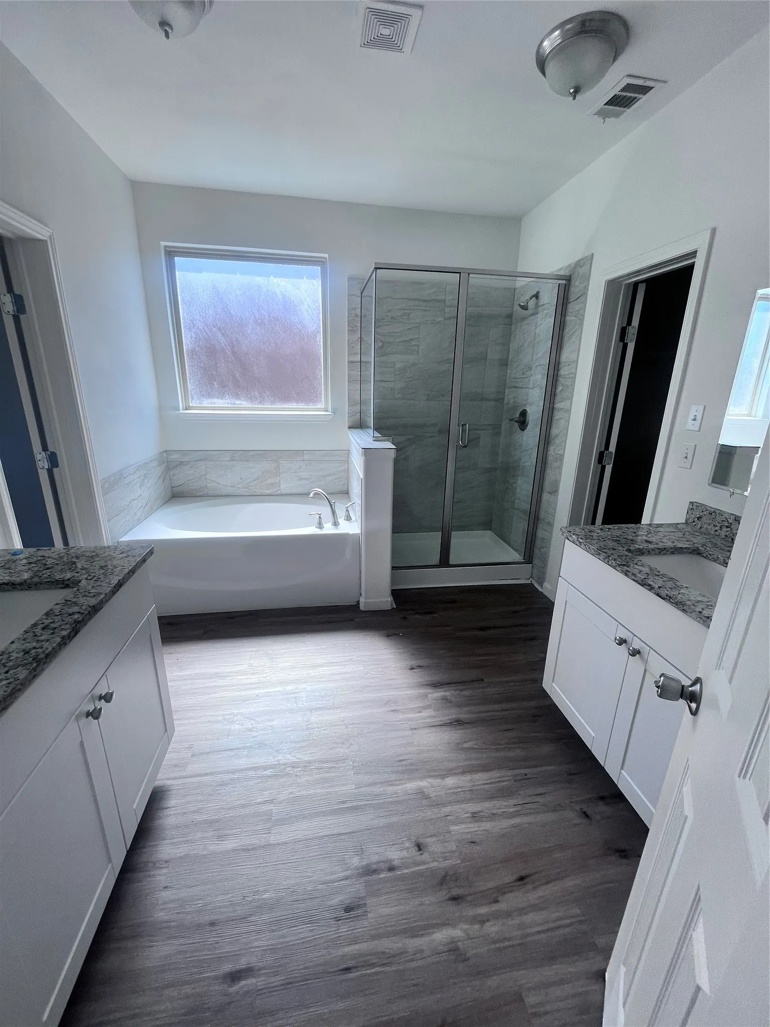 Bathroom featuring a soaking tub, glass-enclosed shower, dual vanity sinks with granite-style countertops, white cabinetry, and wood-finish flooring