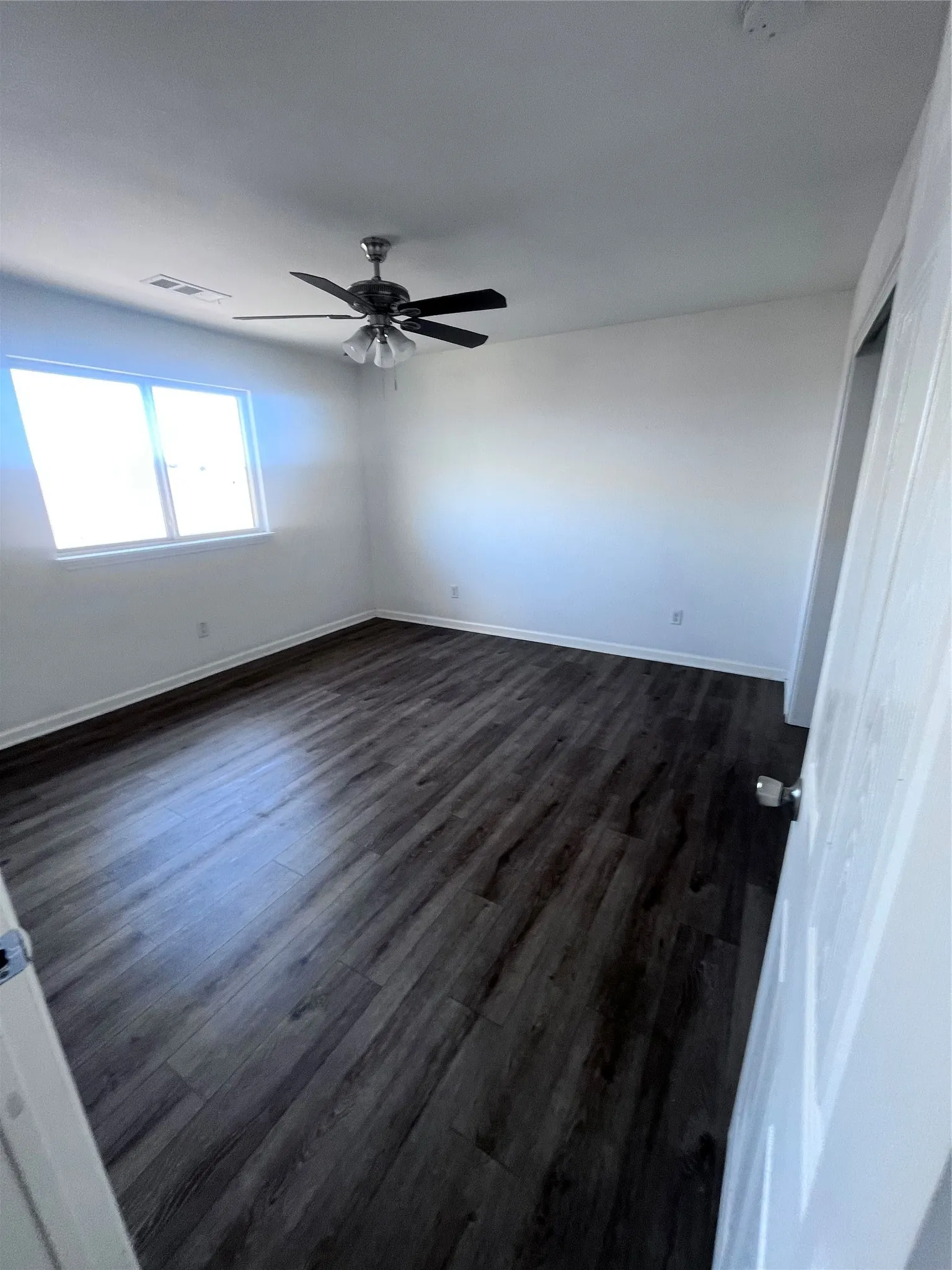 Spacious room featuring dark wood-finish flooring, a ceiling fan with integrated lighting, a large window, and white walls