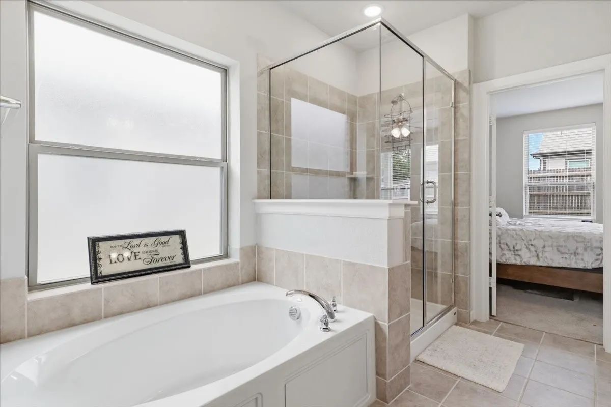White built-in soaking tub with chrome fixtures, large frosted window, and a glass-enclosed shower with tiled walls and a built-in shelf