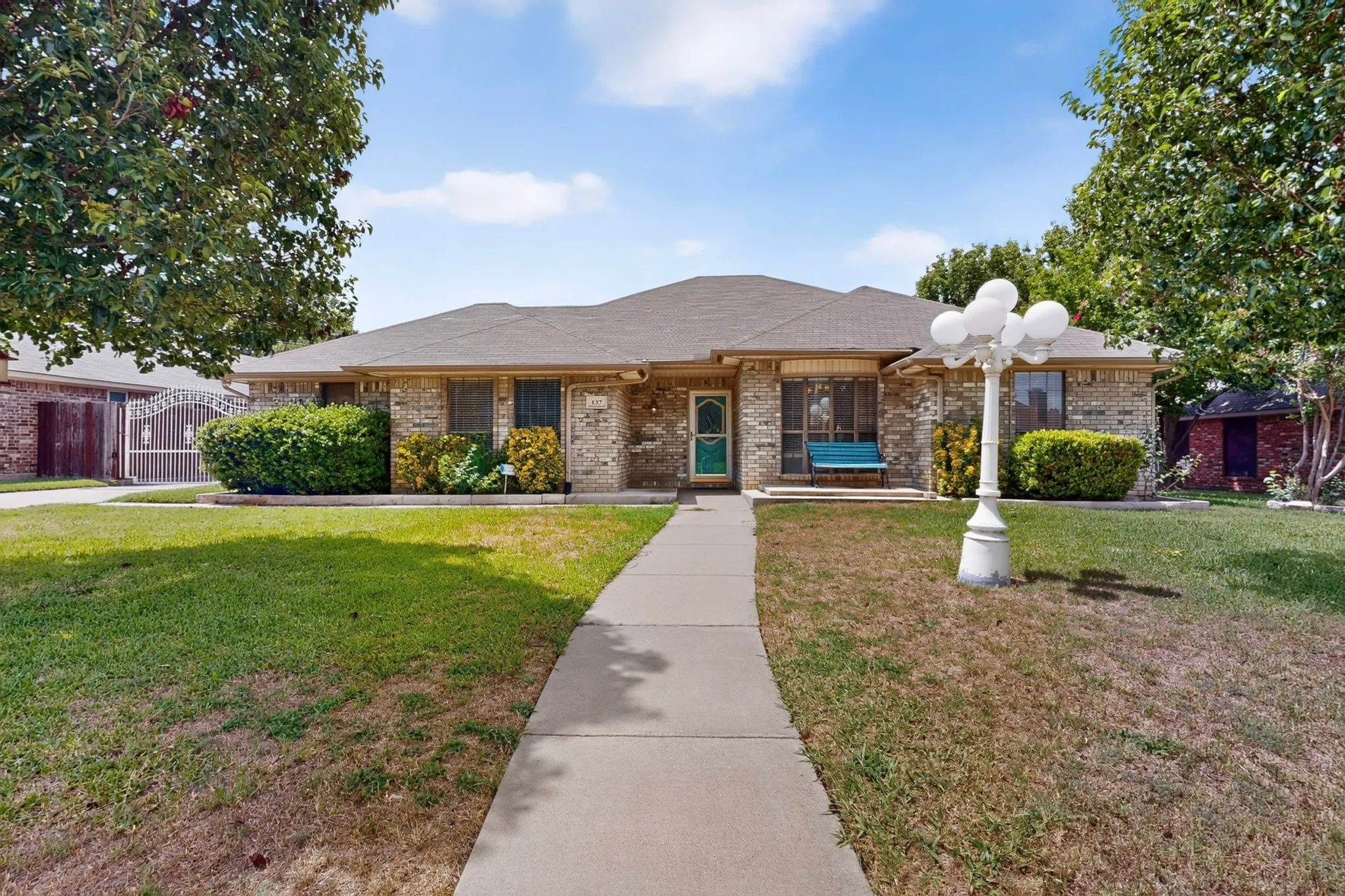 Brick exterior residence featuring a gabled roof, a concrete walkway, mature trees, and established shrubs
