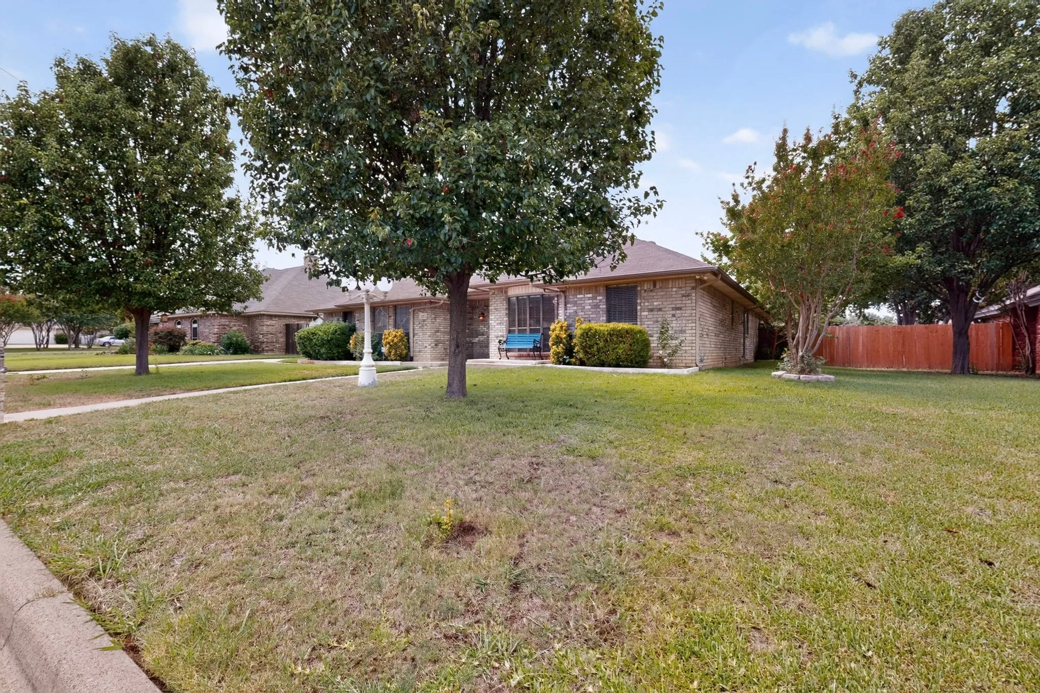 Brick exterior home featuring a gabled roofline, mature trees, a manicured lawn, and a wooden privacy fence