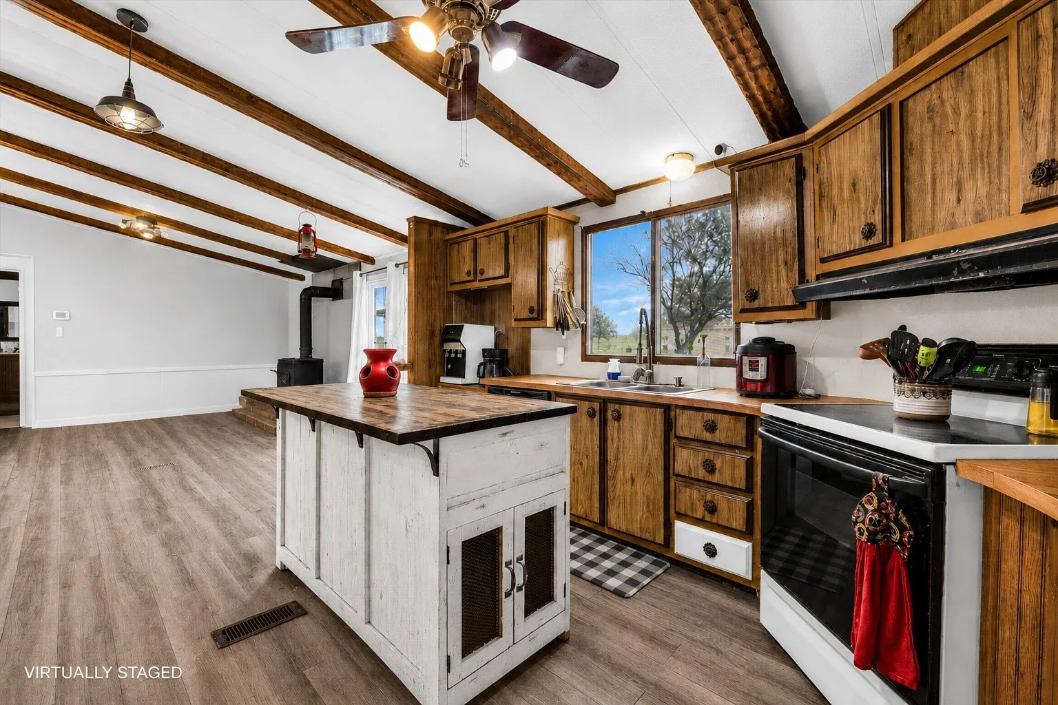 Open-concept kitchen and living area featuring exposed wood ceiling beams, wood-finish flooring, a freestanding wood-burning stove, wood cabinetry, and a white kitchen island.