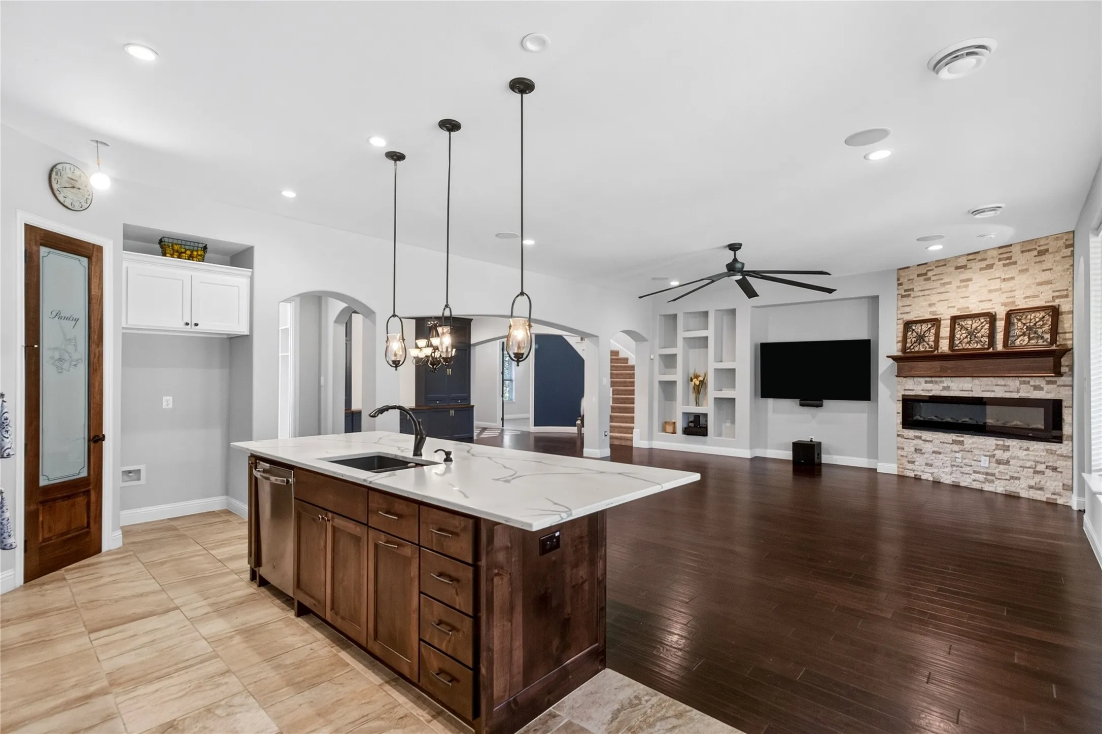 Kitchen island with a white marble-look countertop and integrated sink, pendant lighting, built-in shelving, a stone accent wall, an electric fireplace, and dark wood-finish flooring