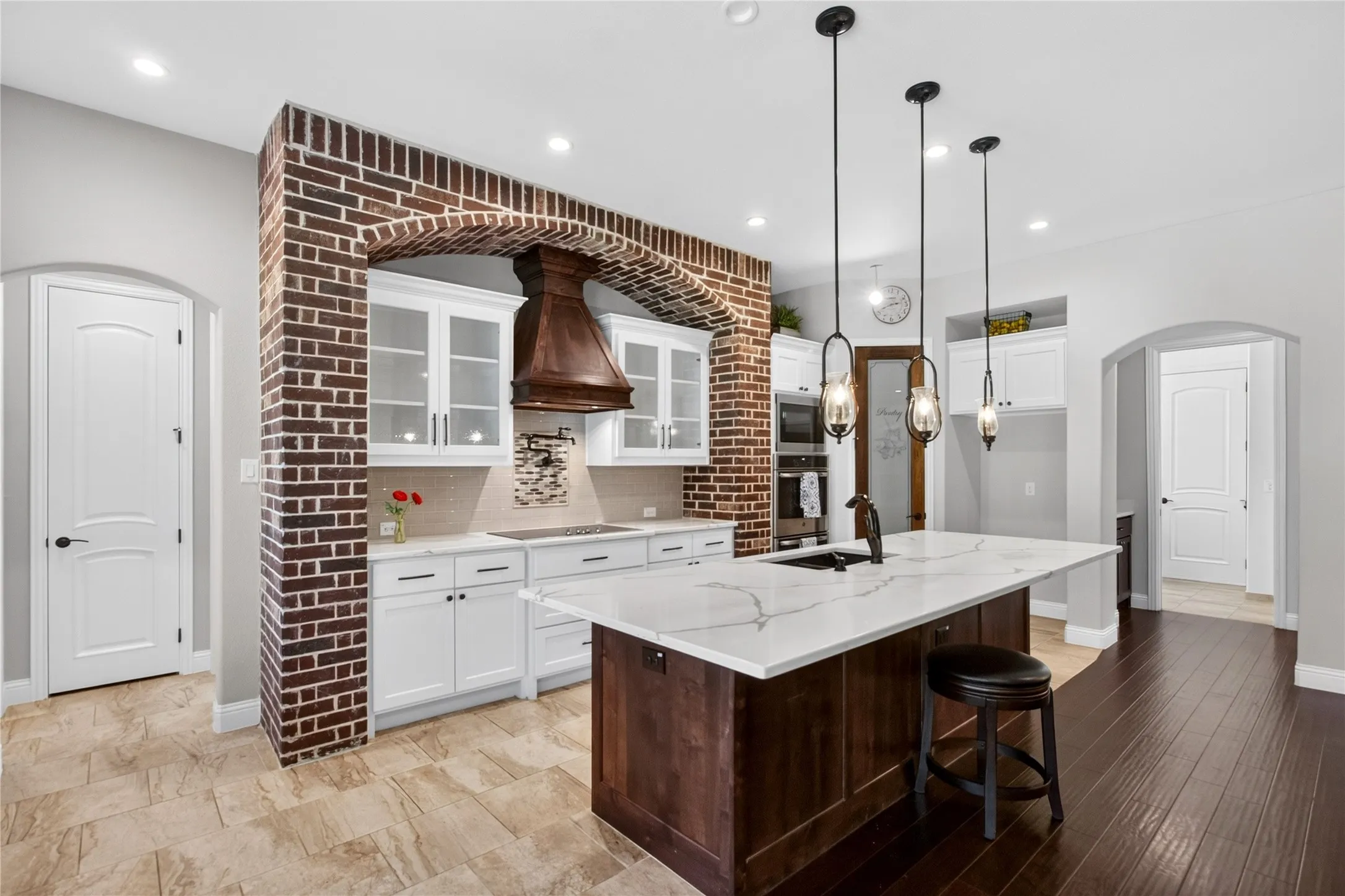 Kitchen featuring a white countertop island with integrated sink and three pendant lights, white cabinetry with glass-front upper units, wood-finish flooring, a brick archway detail, and a wood-finish range hood