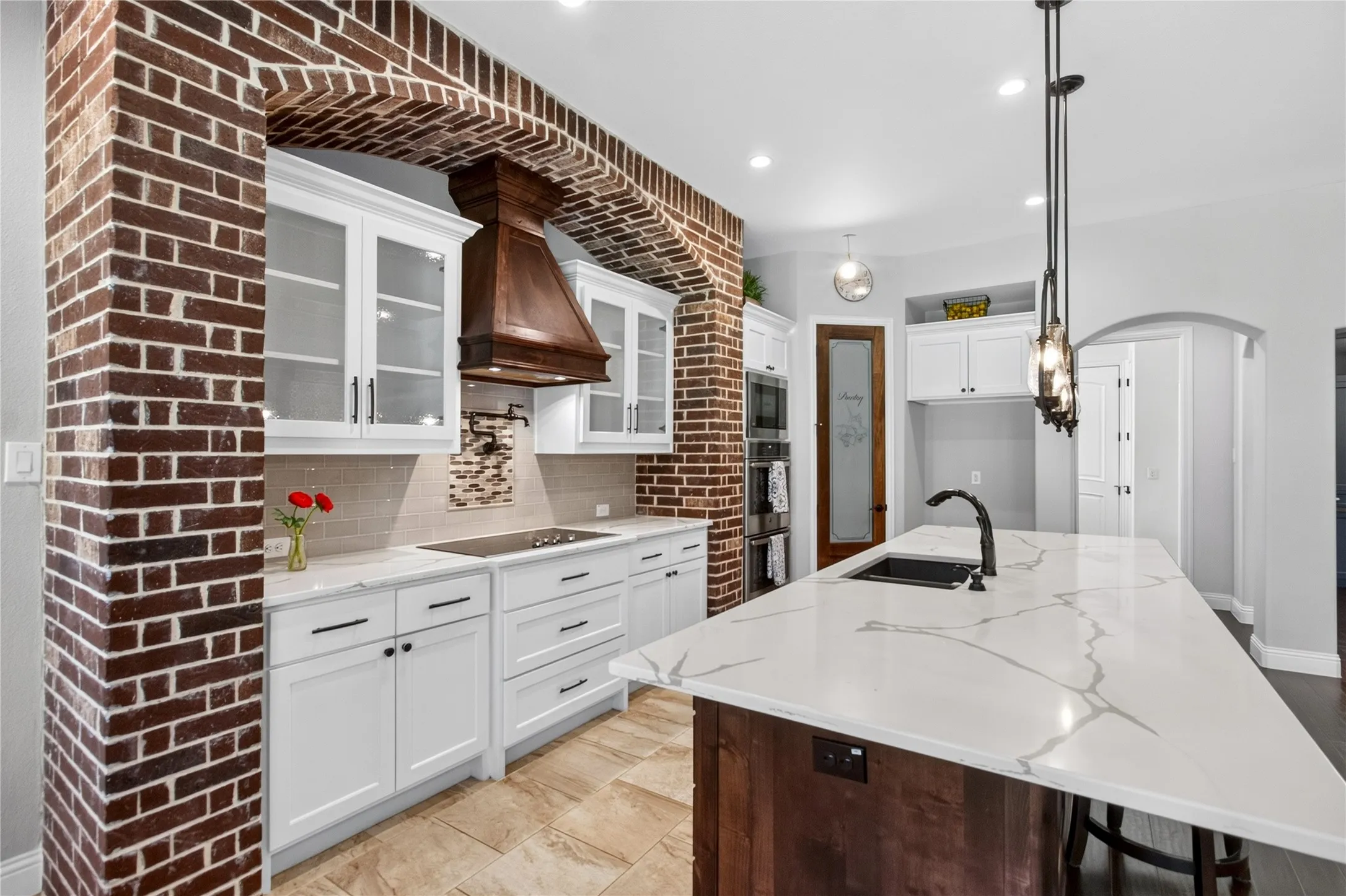 Kitchen featuring a brick archway, white cabinetry, a wood-finish range hood, light-toned tile flooring, and a central island with a waterfall countertop