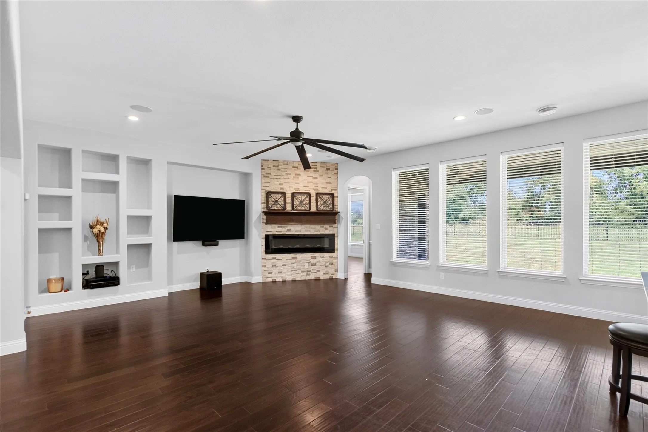 Spacious living area featuring rich wood-finish flooring, a contemporary ceiling fan, and a stacked stone fireplace with a wood mantle