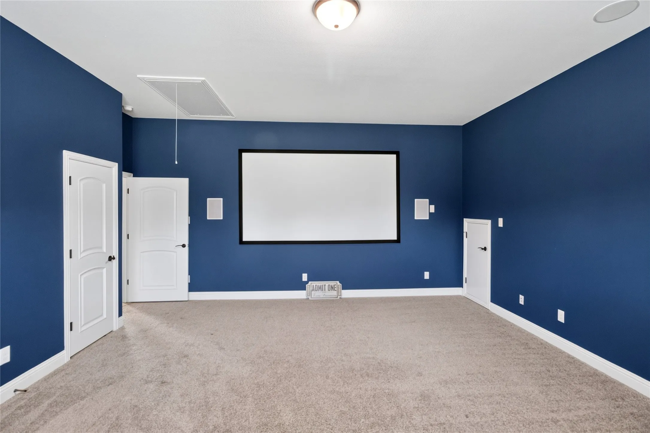 Dedicated media room featuring a large projection screen, built-in speakers, plush carpet flooring, and a ceiling-mounted light fixture