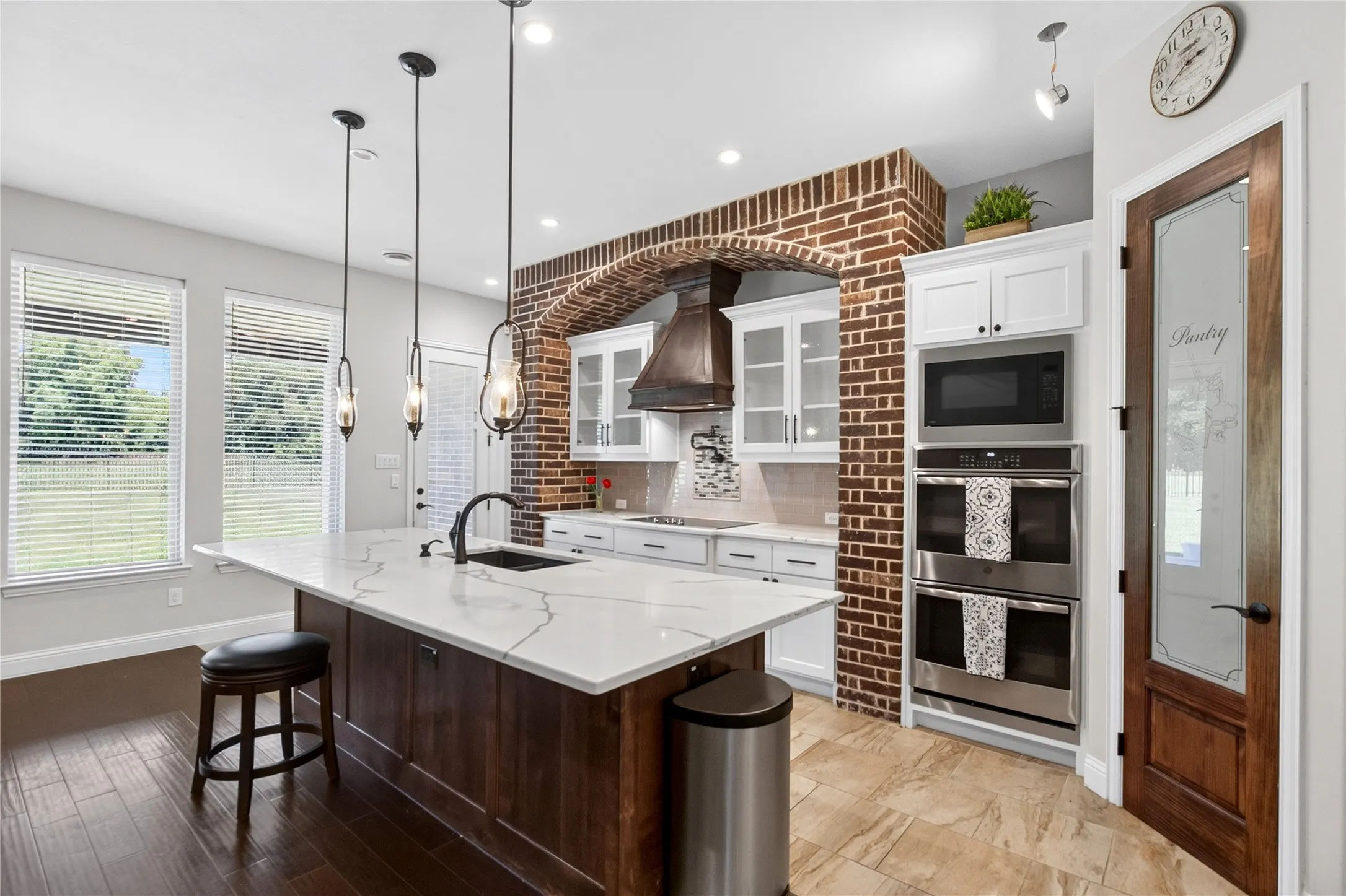 Gourmet kitchen featuring a large island with white stone-finish countertop, dark wood-finish cabinetry, stainless steel double ovens, and a brick archway