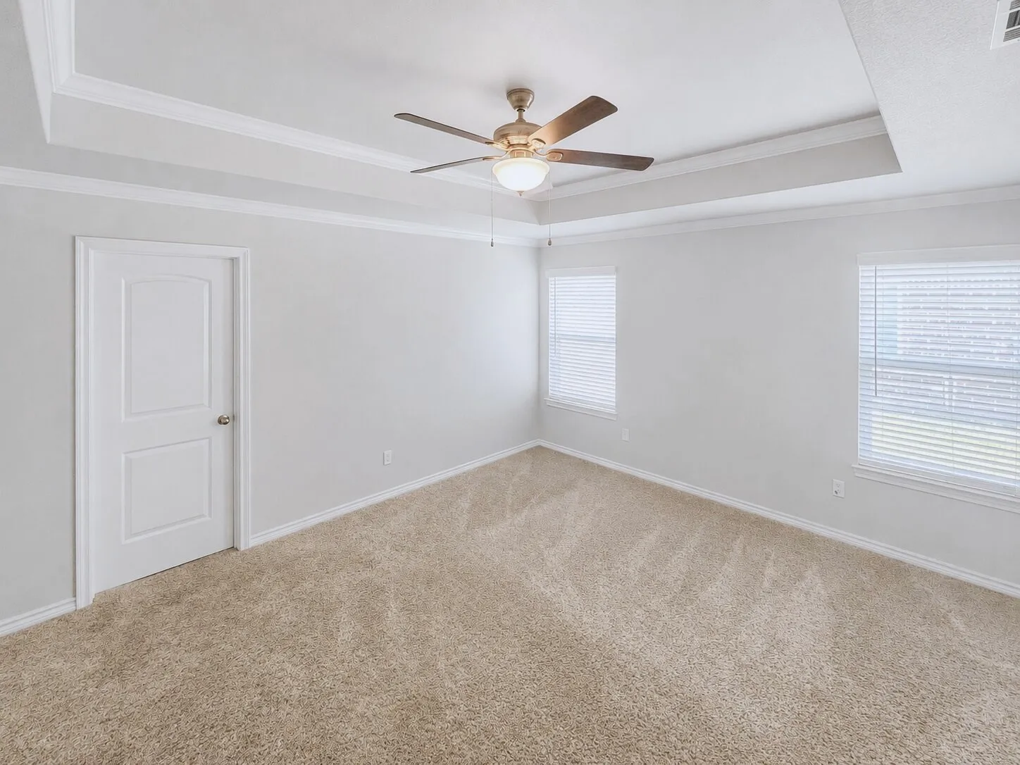 Spacious room featuring tray ceilings with crown molding, a ceiling fan with integrated lighting, two windows with blinds, and light-toned carpeting