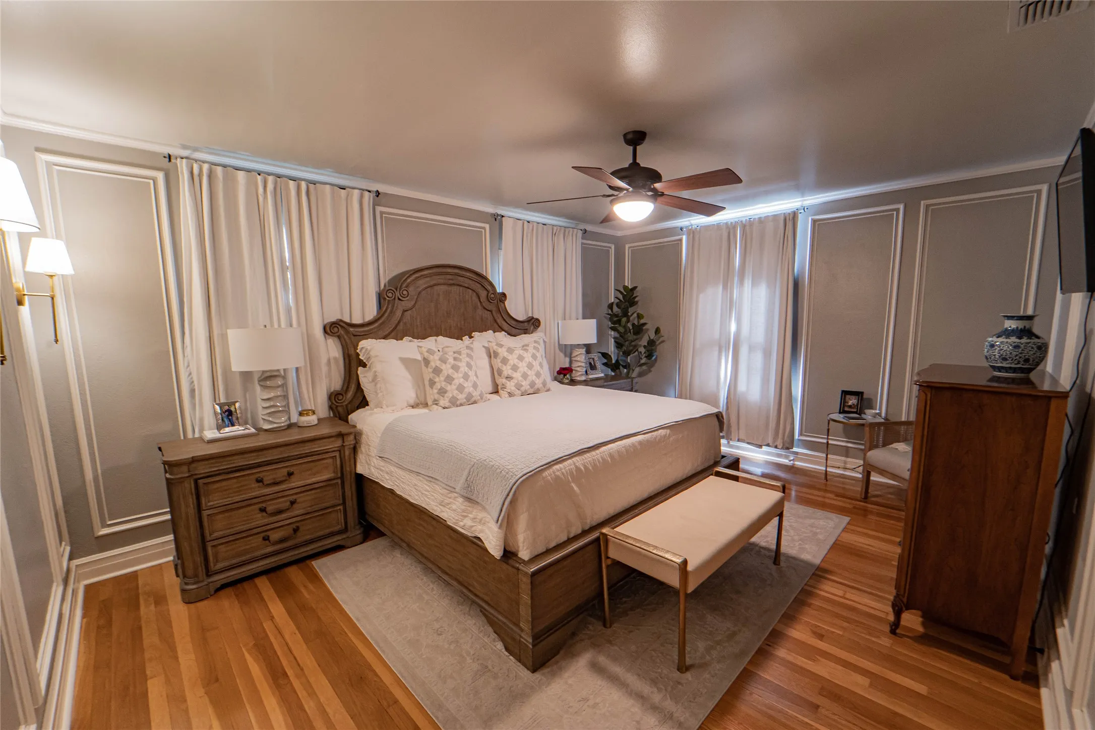 Bedroom featuring hardwood flooring, decorative wall molding, ceiling fan with light fixture, and window treatments
