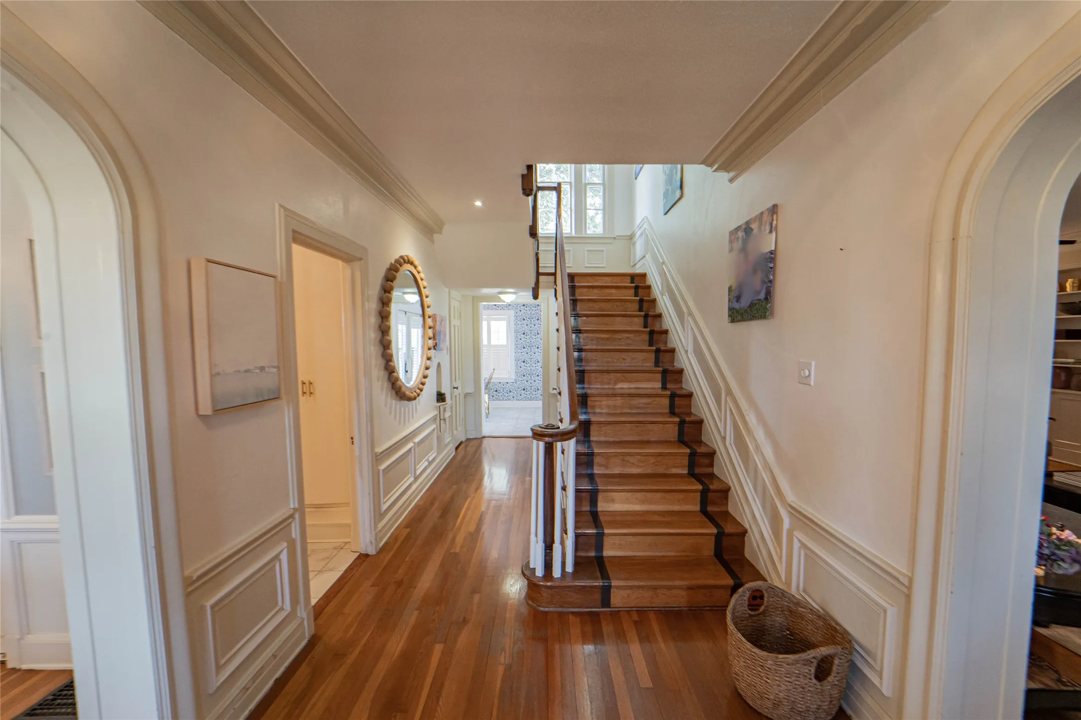 Grand foyer featuring wood-finish flooring, a prominent staircase with wood treads and handrail, wainscoting, arched doorways, and crown molding