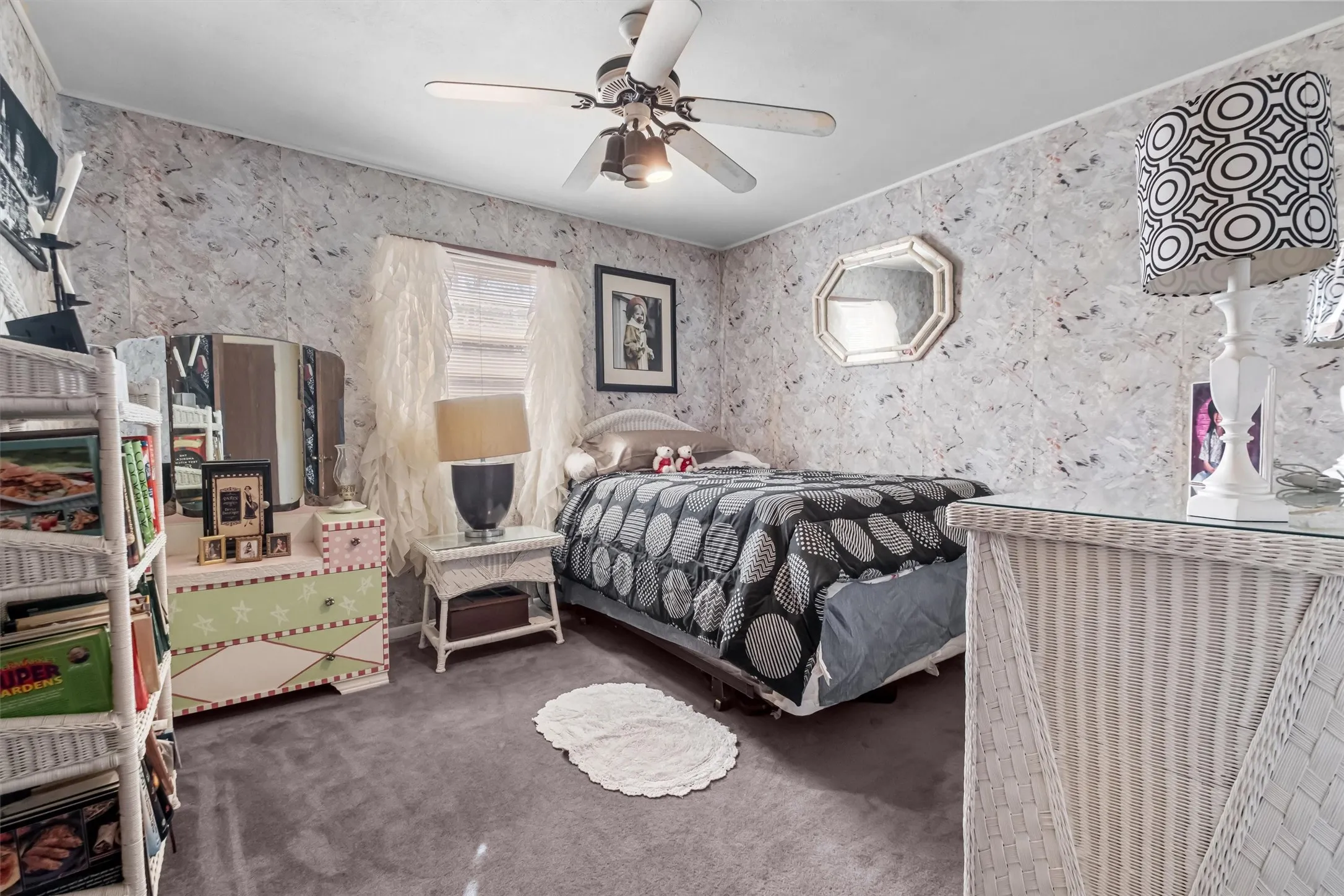 Carpeted room featuring a ceiling fan with integrated lighting, a window with sheer curtains, textured wallpaper, and an octagonal wall mirror