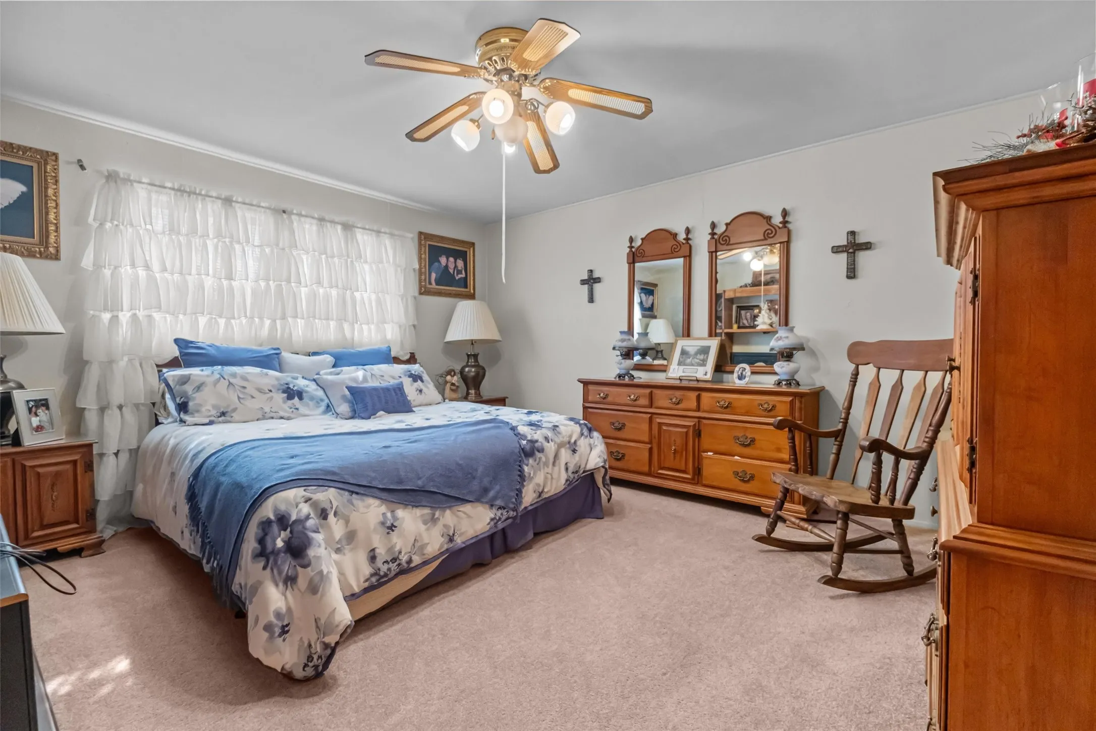 Neutral-toned carpeting extends throughout this room, featuring a ceiling fan with light fixture, white crown molding, and a spacious window with layered white curtains