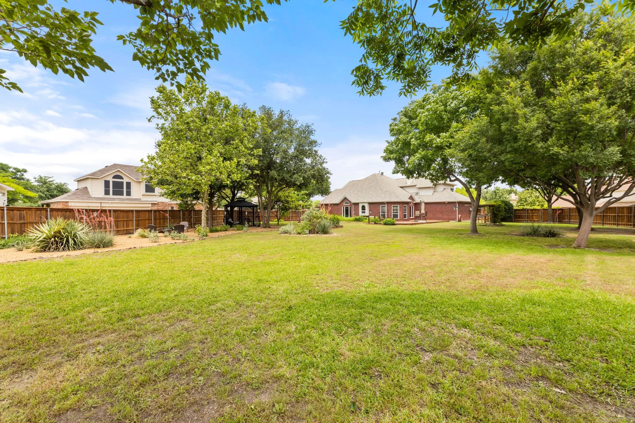 Expansive backyard featuring a large lawn area, mature trees, a wooden privacy fence, a detached gazebo, and landscaped garden beds