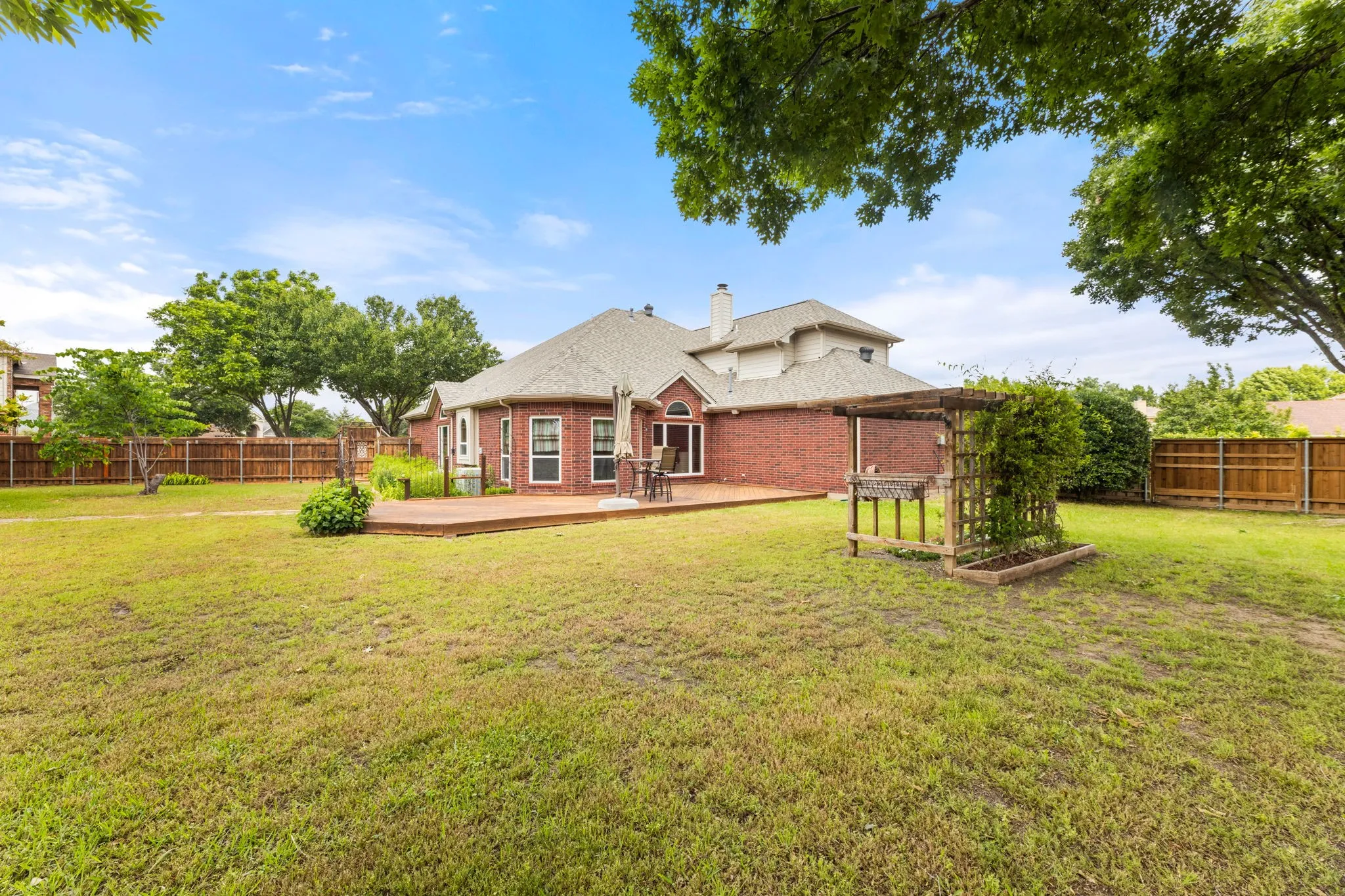 Spacious lawn featuring a brick exterior, a multi-level wood deck, an arched window, and a wooden pergola with a built-in planter