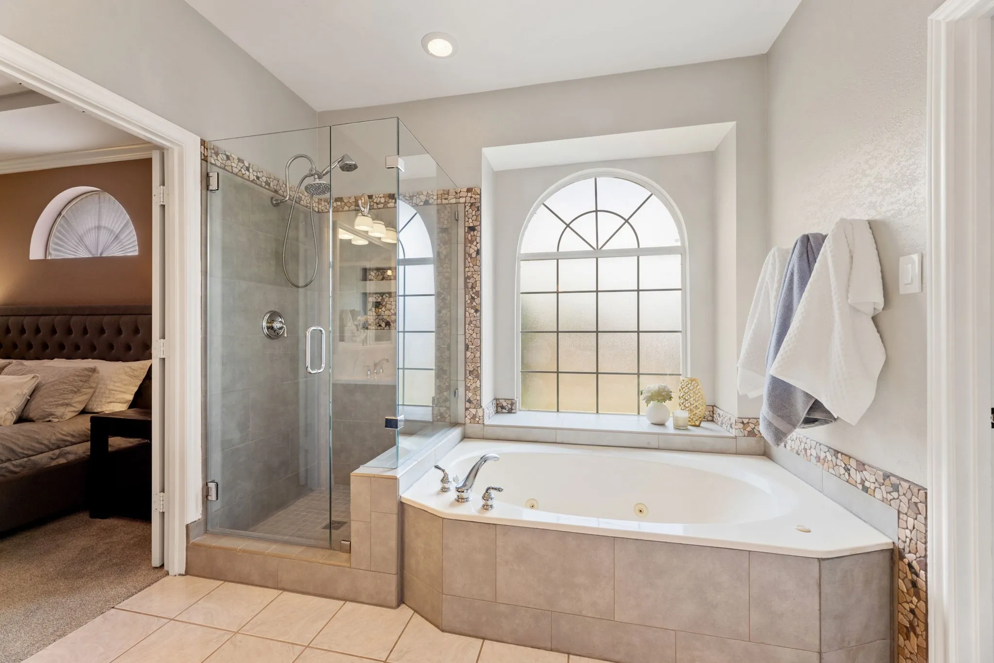 Corner soaking tub with a tiled surround, arched window, and mosaic tile accent