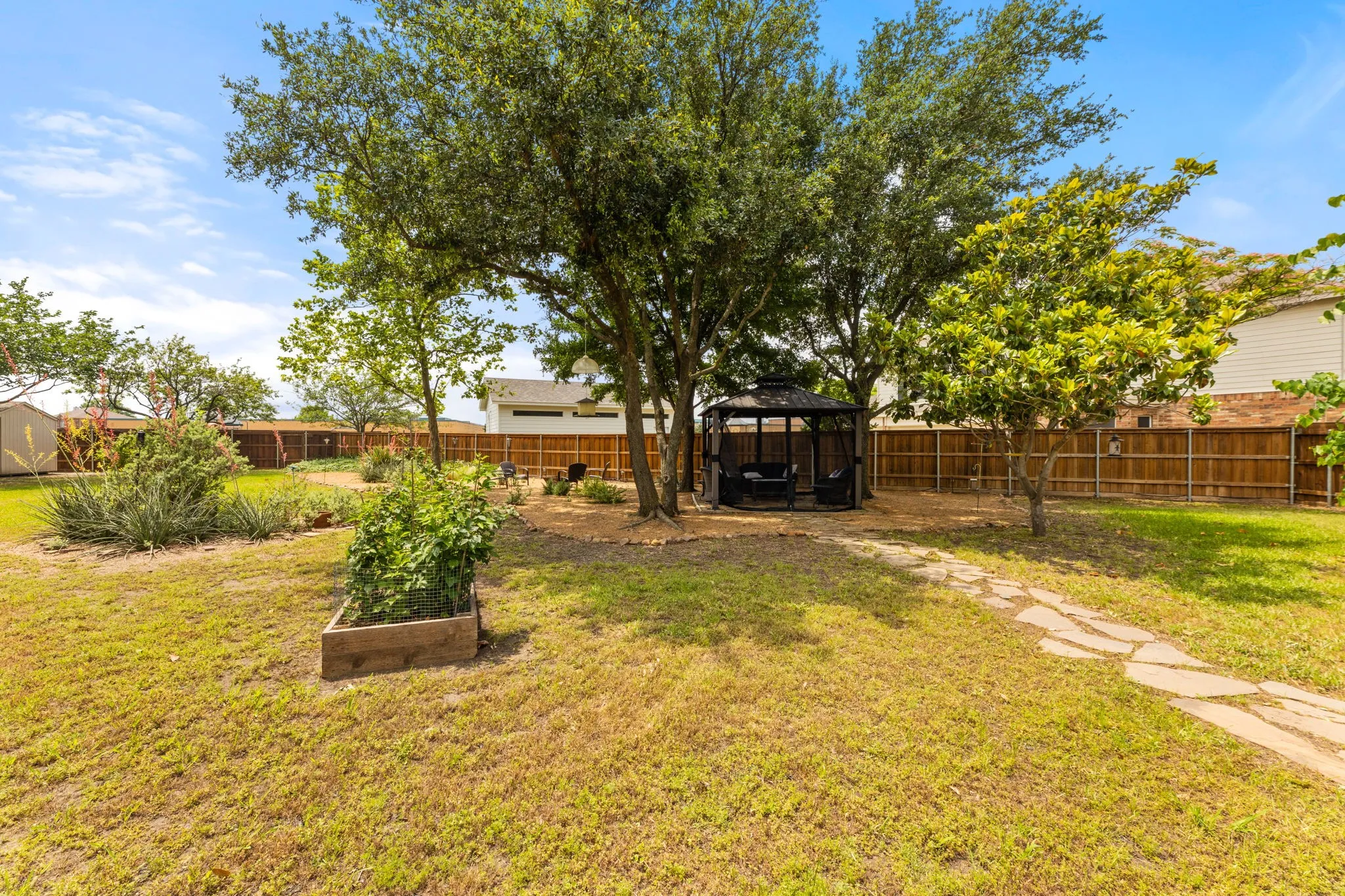 Expansive backyard featuring mature trees, a stone pathway, a dark-framed gazebo, a wooden privacy fence, and a raised garden bed