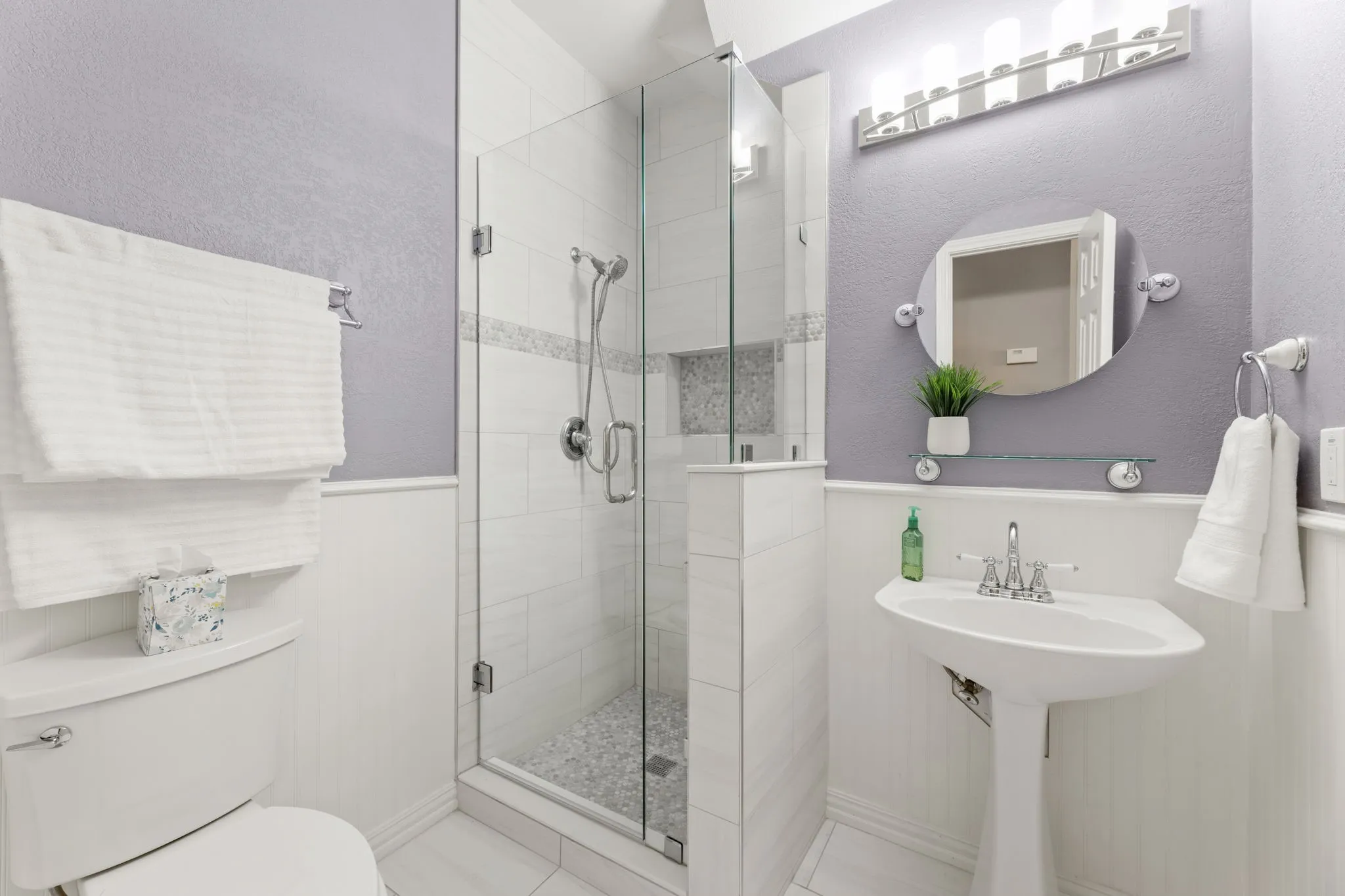 Full bathroom featuring a glass-enclosed shower with pebble-finish flooring, a white pedestal sink, and a circular wall mirror