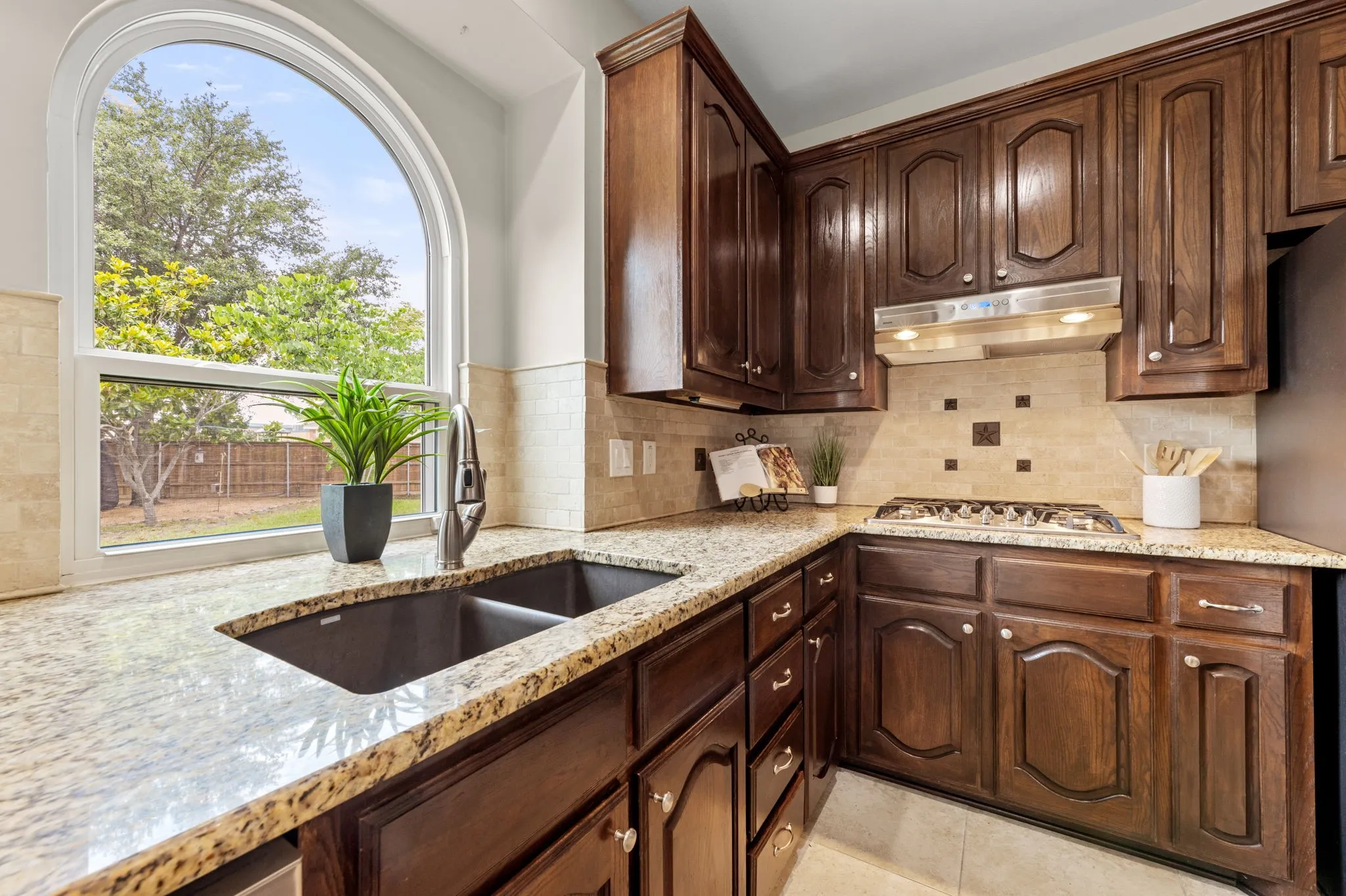 Kitchen featuring a prominent arched window, granite countertops, dark wood cabinetry, a double basin sink, and a tile backsplash with decorative accents