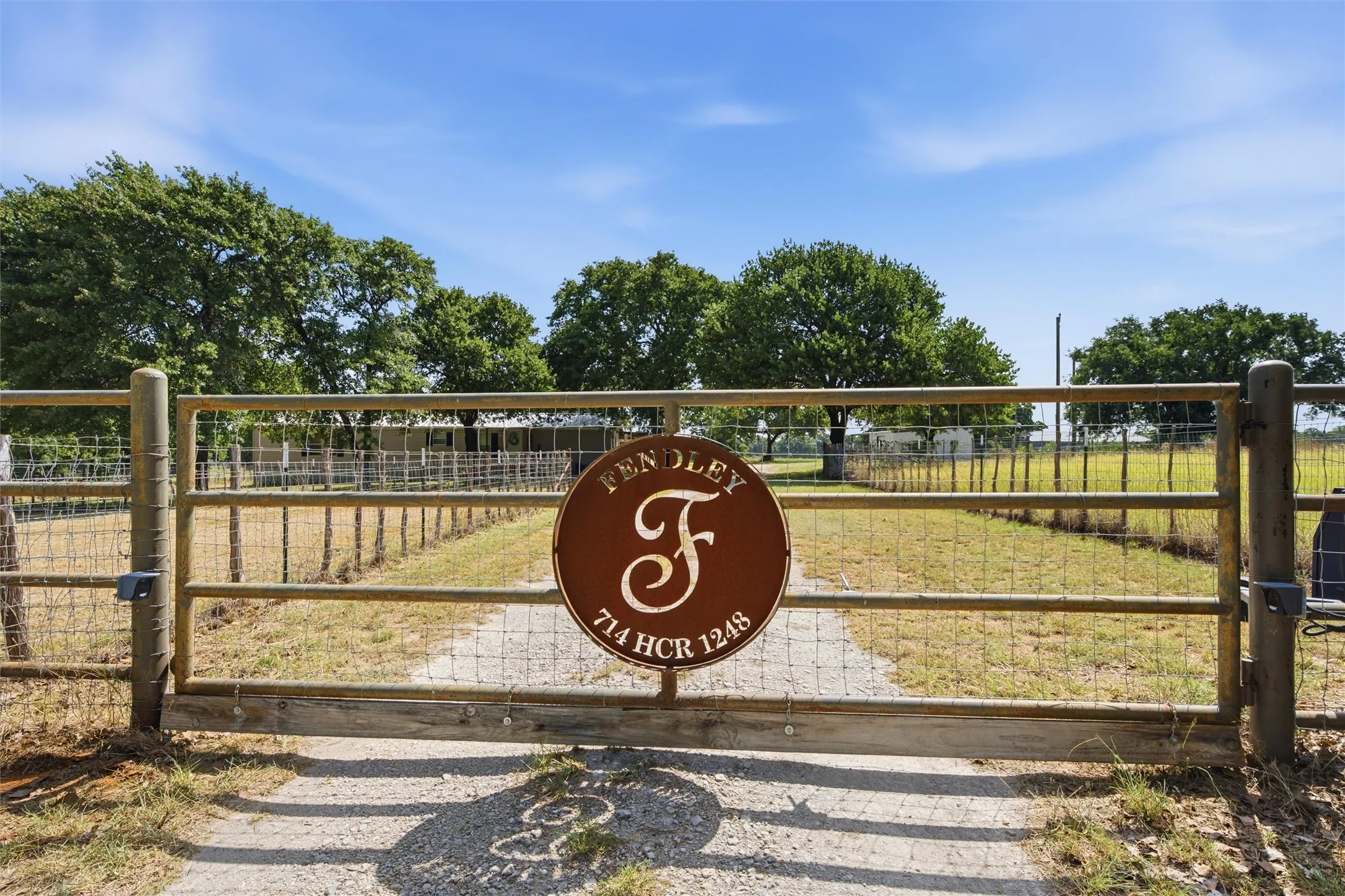 Gravel driveway leading to a metal gate with wire fencing, featuring mature trees and open land