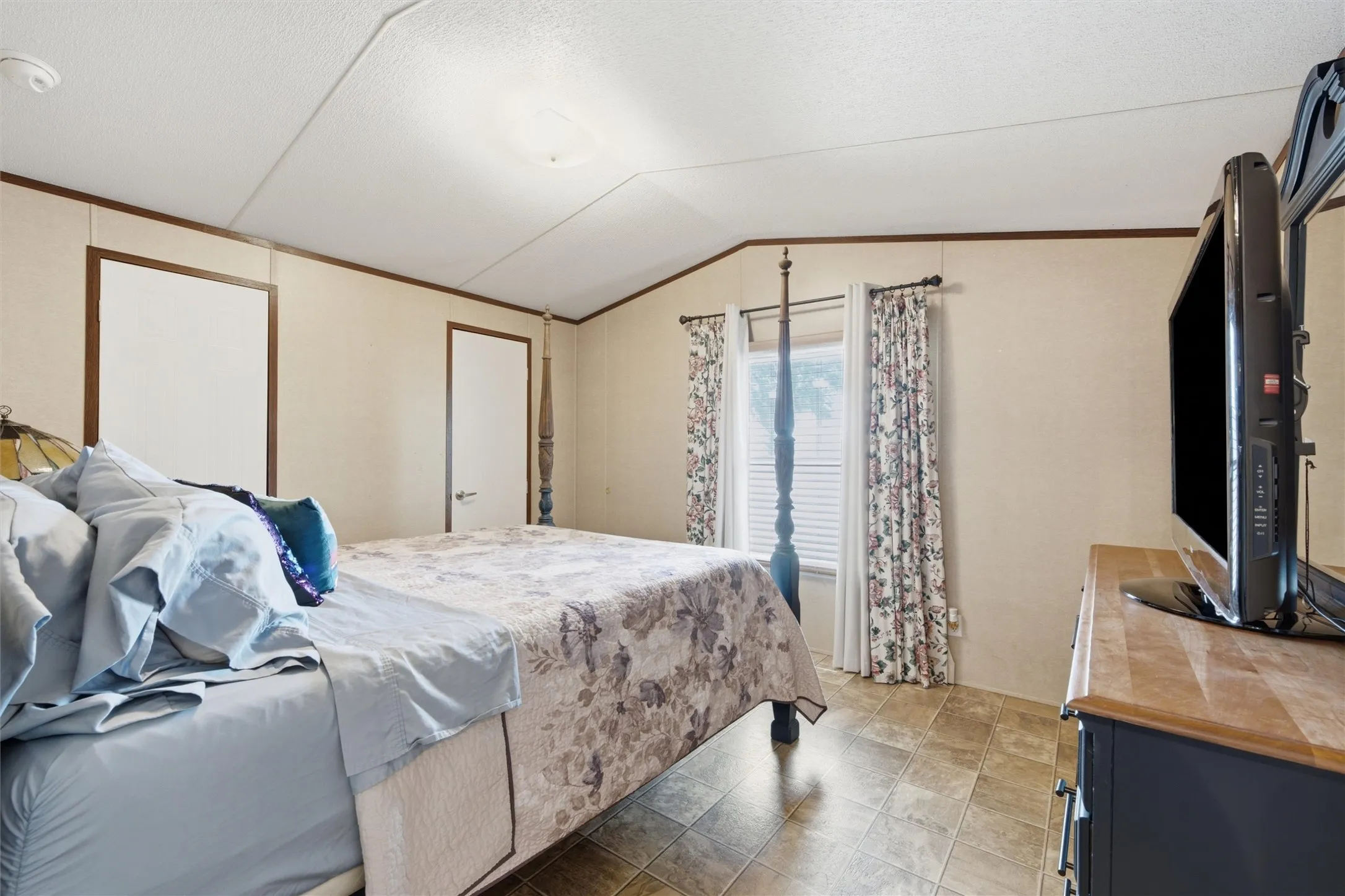 Spacious bedroom featuring light-toned walls with dark trim, durable tile-finish flooring, a vaulted ceiling, and dual windows with blinds
