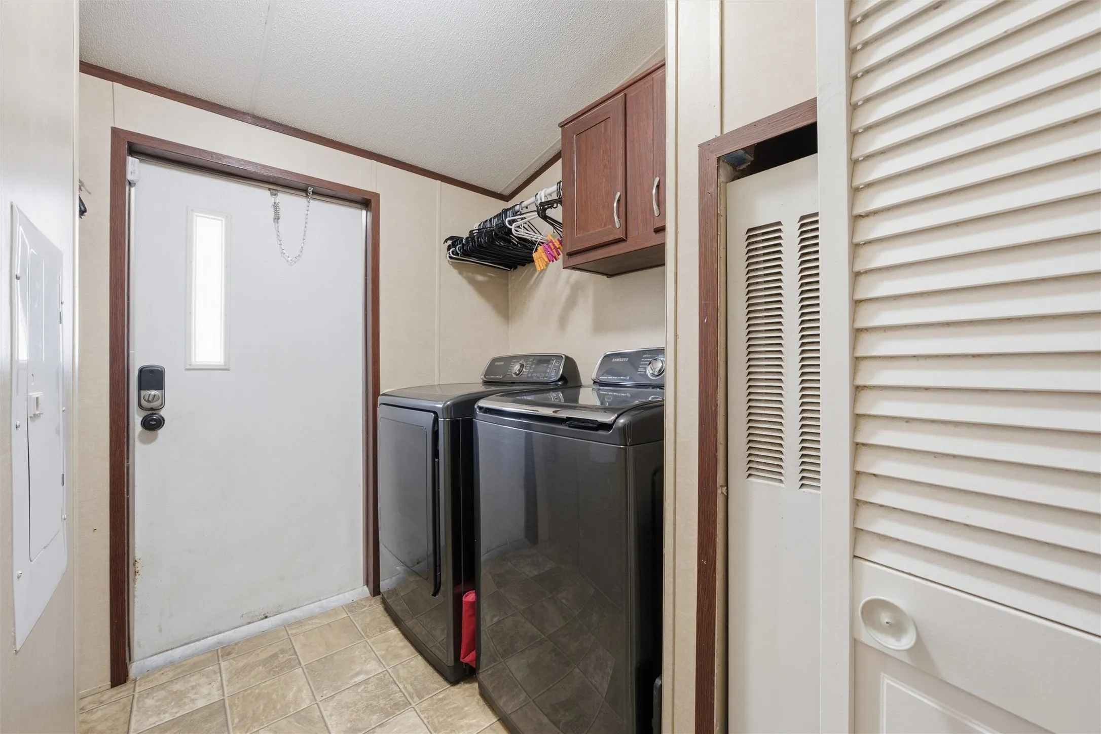 Dedicated laundry area featuring a white entry door with a vertical glass insert, modern dark appliances, overhead wood-finish cabinetry, a built-in wire rack, and tiled flooring