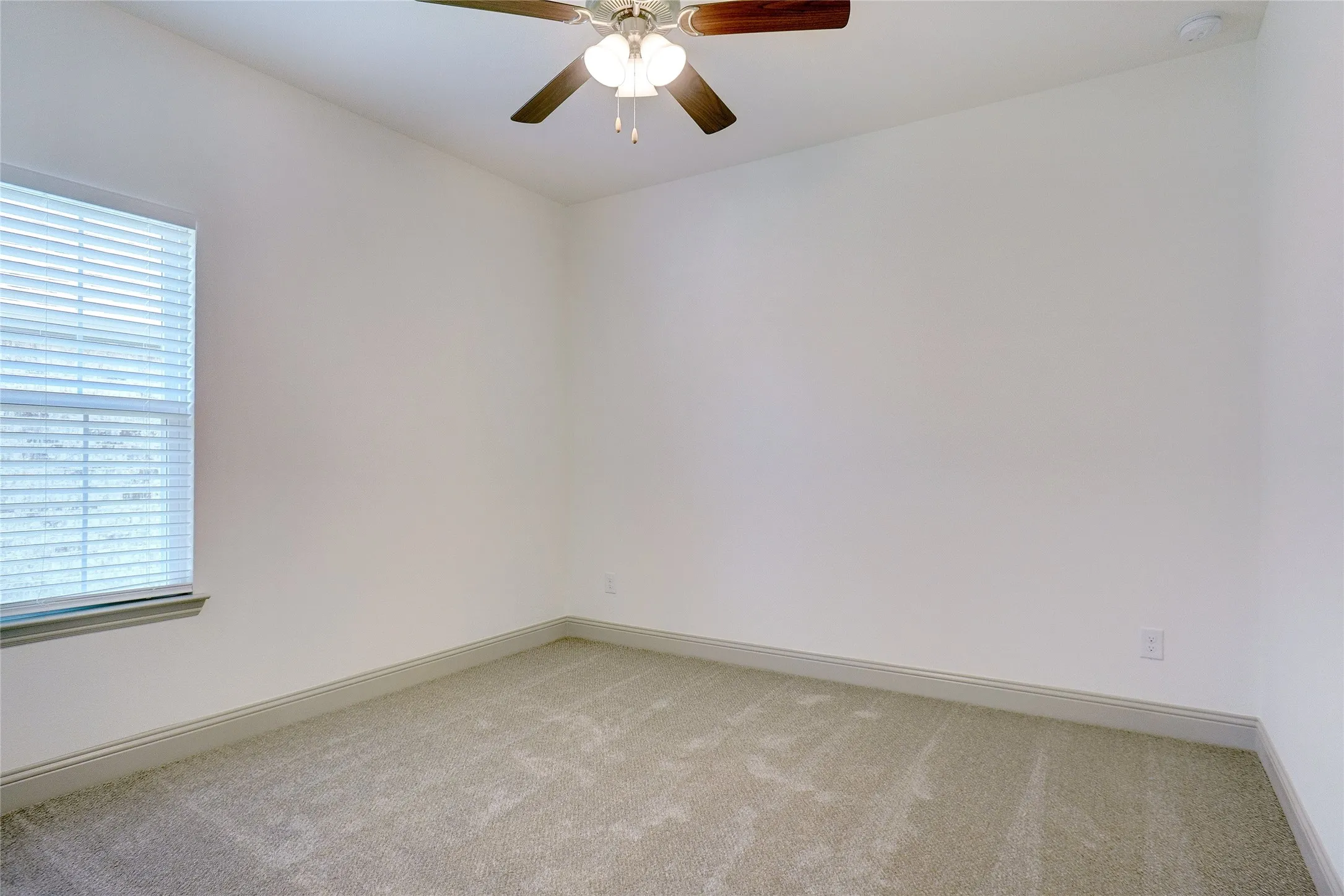 Carpeted room with a ceiling fan featuring a central light fixture, a single window with horizontal blinds, and white baseboards