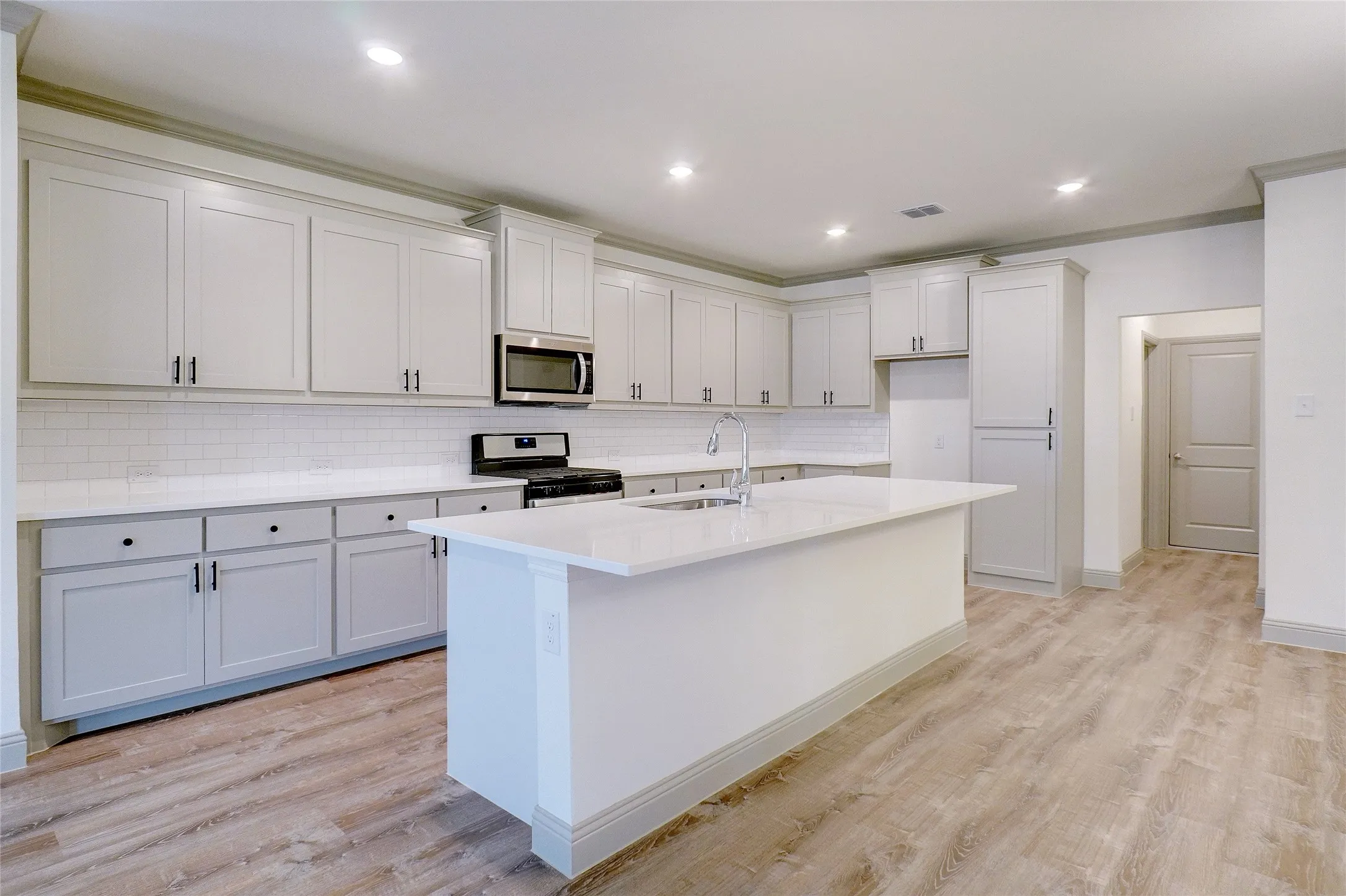 Contemporary kitchen featuring light wood-finish flooring, white shaker-style cabinetry with dark hardware, subway tile backsplash, and light-toned countertops