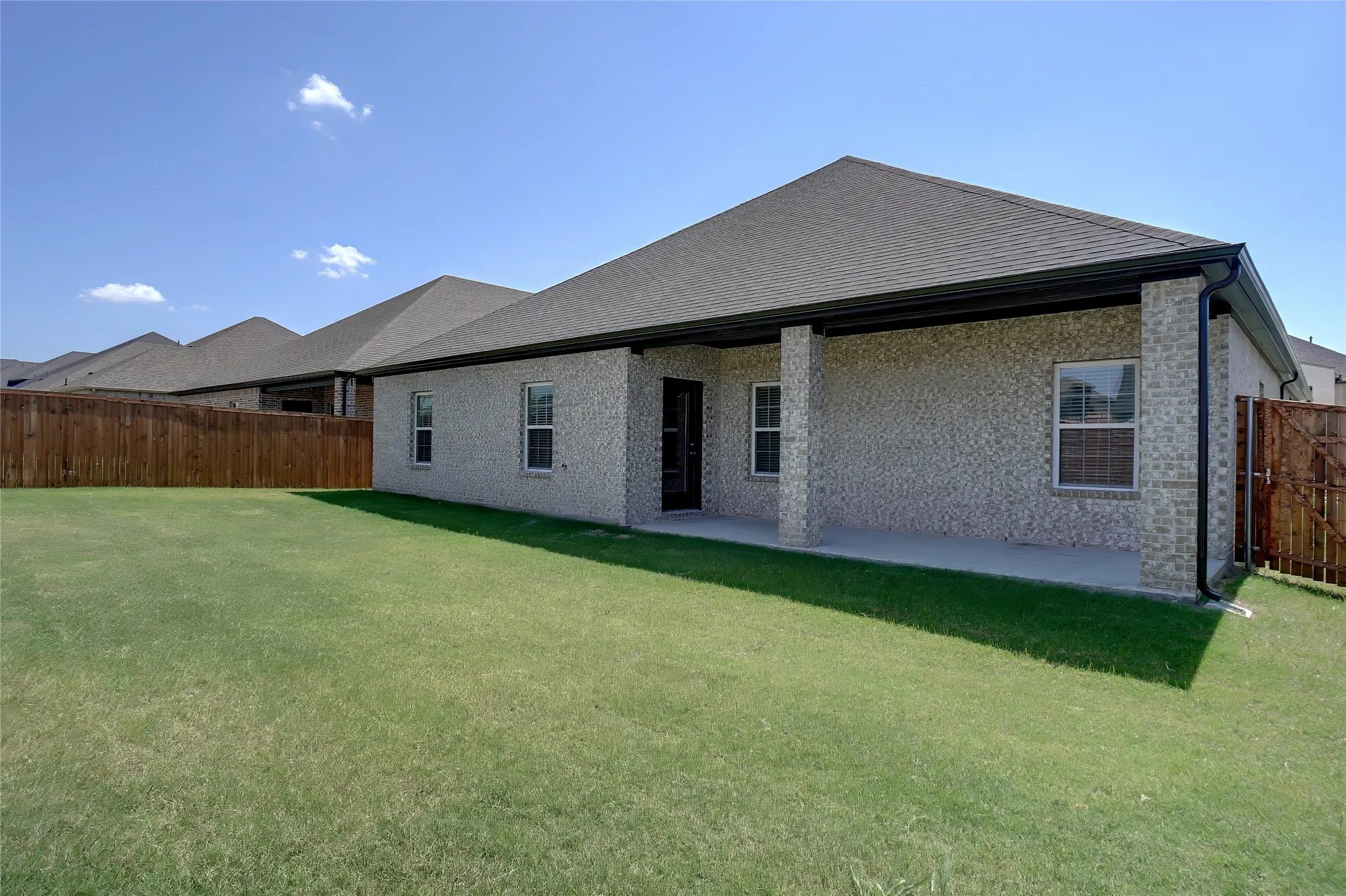 Expansive green lawn with a covered patio, light-colored brick exterior, dark shingle roof, and a privacy fence