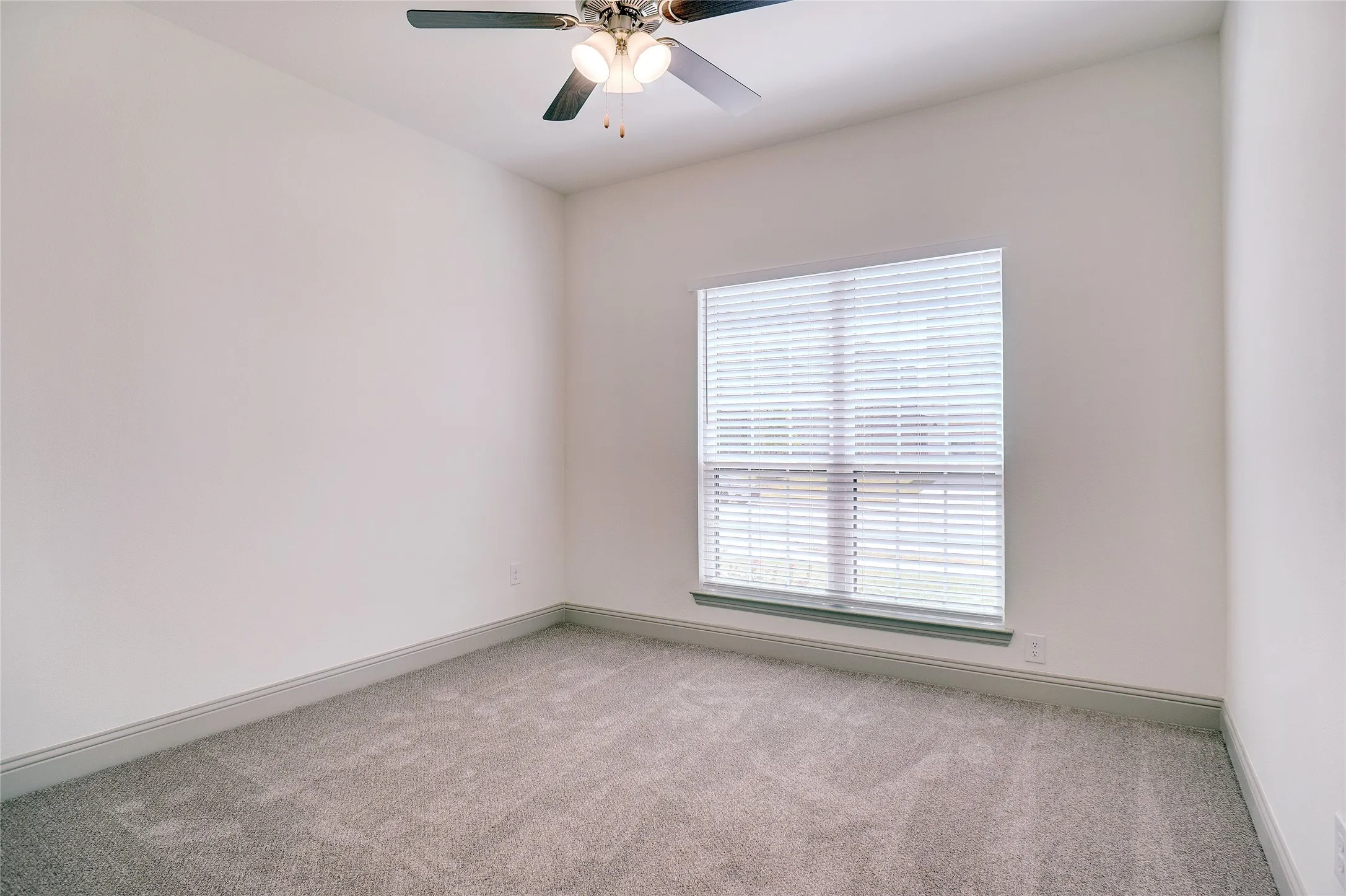 Interior room featuring light gray carpet flooring, white walls, a single window with horizontal blinds, a dark-bladed ceiling fan, and gray baseboard molding