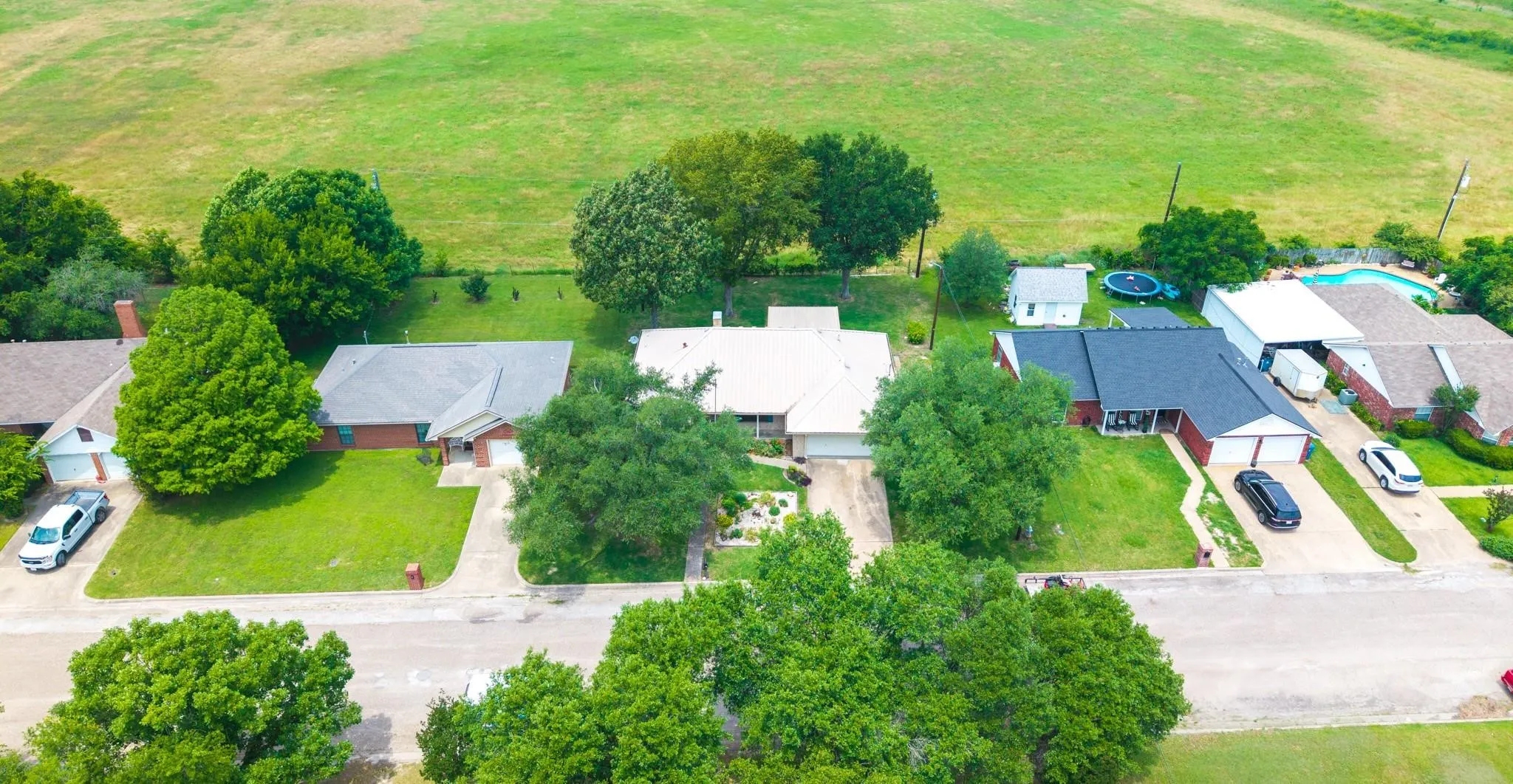 Aerial view of a residential property with a green lawn and mature trees, featuring a light-colored roof and a concrete driveway