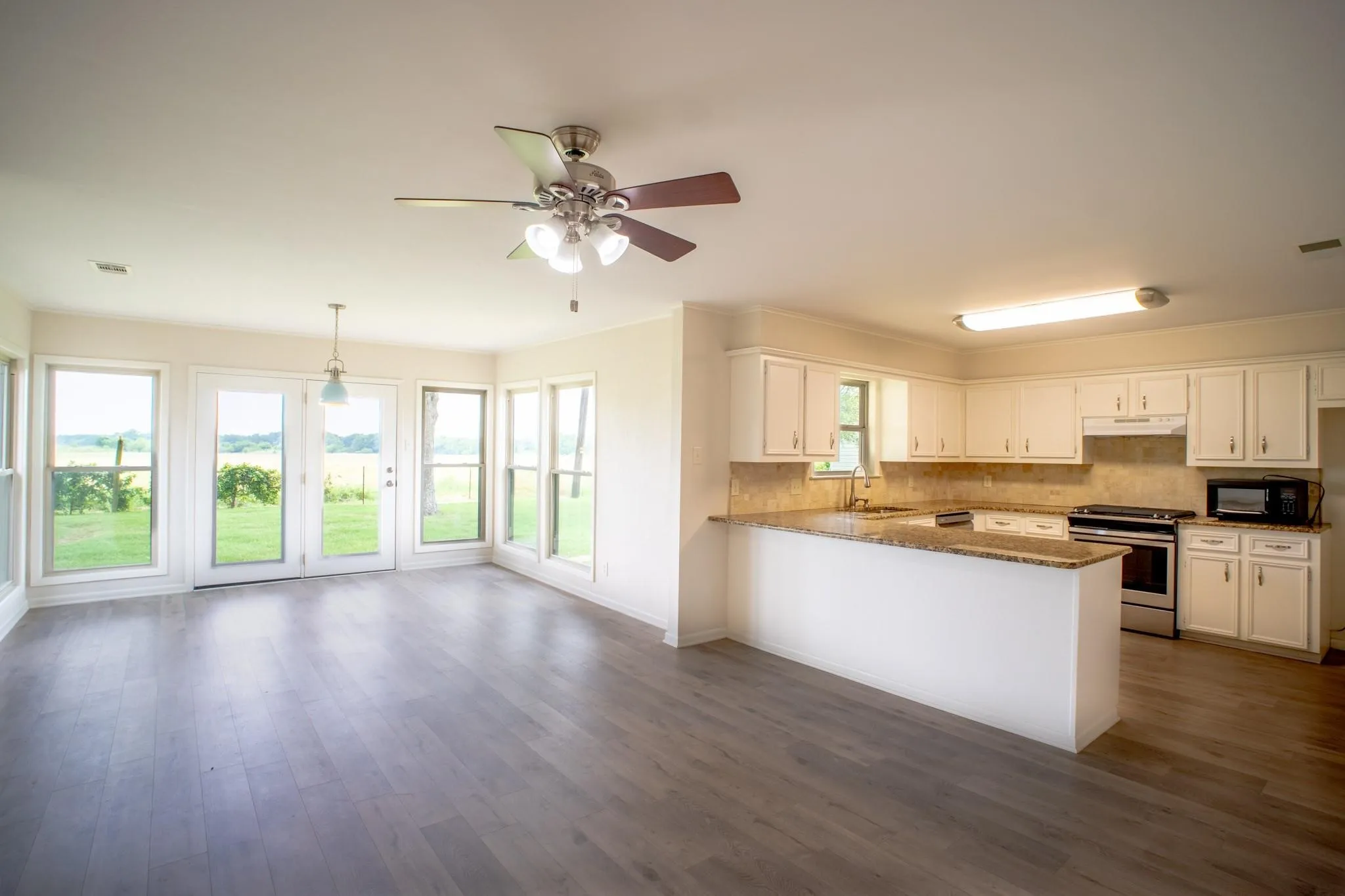 Open-concept living space featuring wood-finish flooring, a kitchen with white cabinetry and granite-style countertops, stainless steel appliances, and a dining area with multiple windows and double glass doors