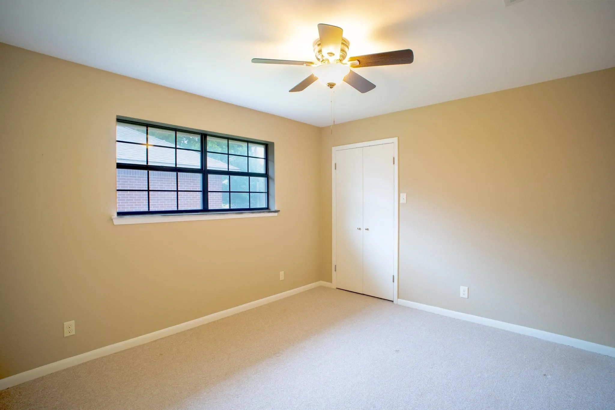 Neutral-toned room featuring a large window with black grilles, a ceiling fan with integrated lighting, carpeted flooring, and a bifold closet door