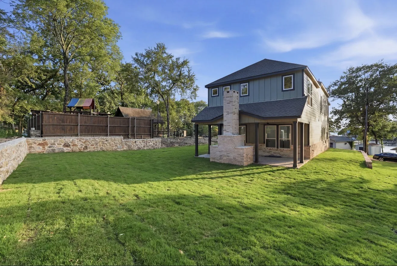 Expansive backyard featuring a stone retaining wall, manicured lawn, and a two-story residence with board and batten siding, multiple windows, and a stone outdoor fireplace with a covered patio