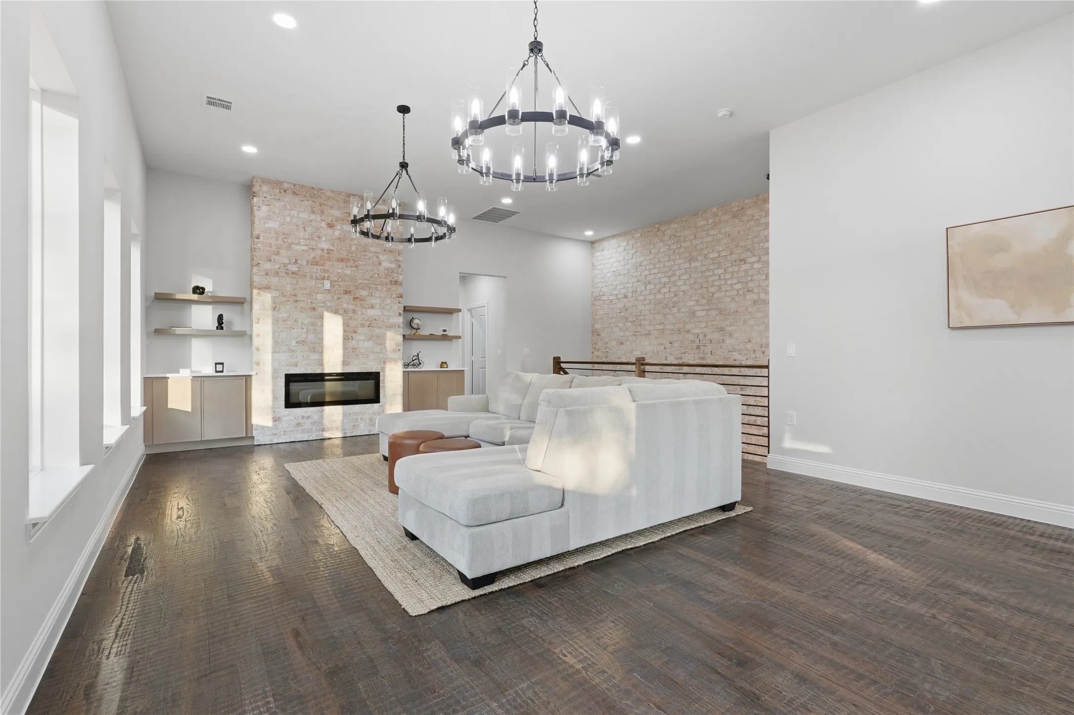 Spacious living area featuring a brick accent wall with a linear fireplace, custom built-in shelving, dark wood-finish flooring, and two contemporary chandeliers