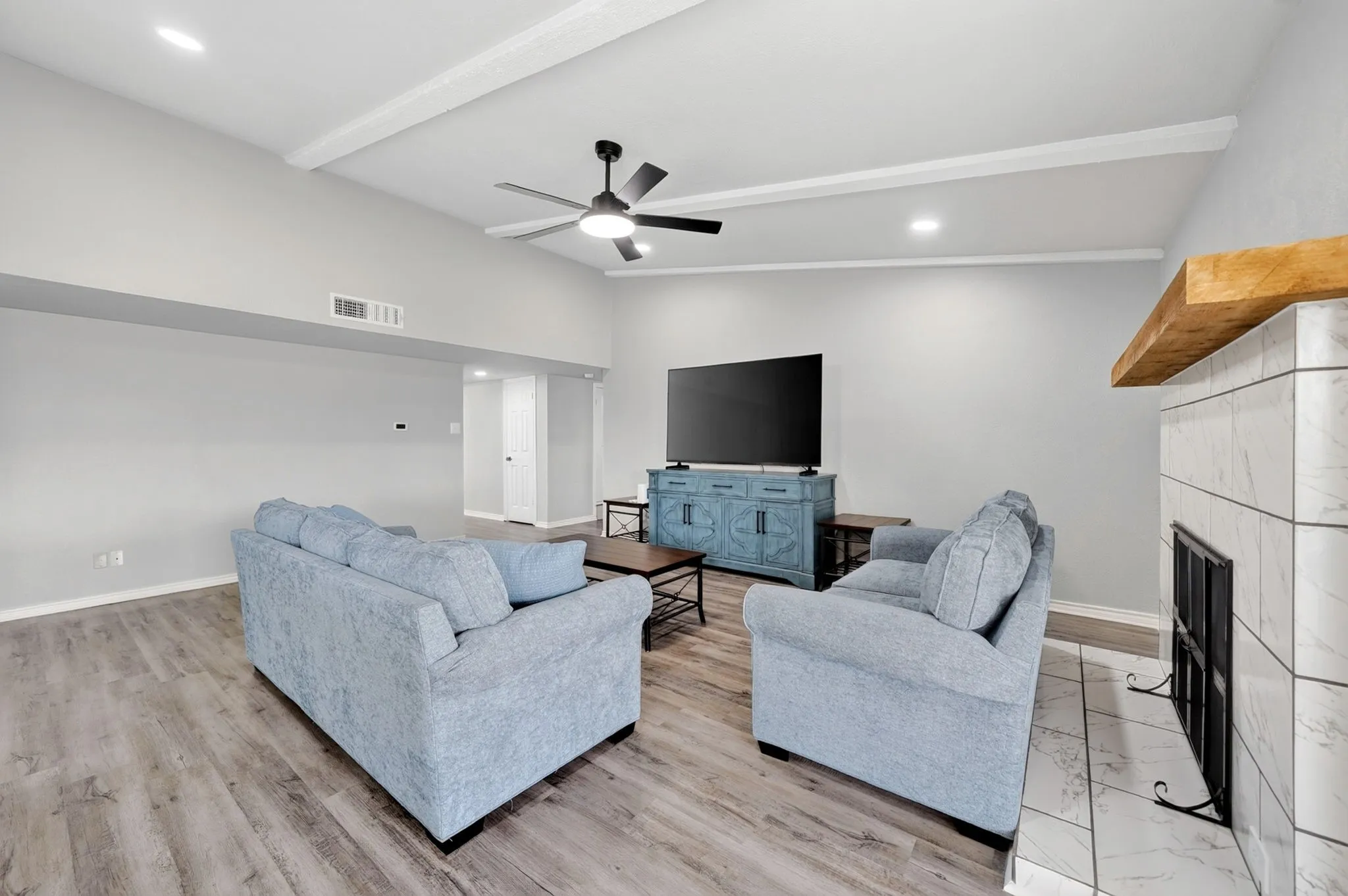 Living area featuring wood-finish flooring, a tiled fireplace with a wood mantle, a vaulted ceiling with exposed beams, recessed lighting, and a ceiling fan